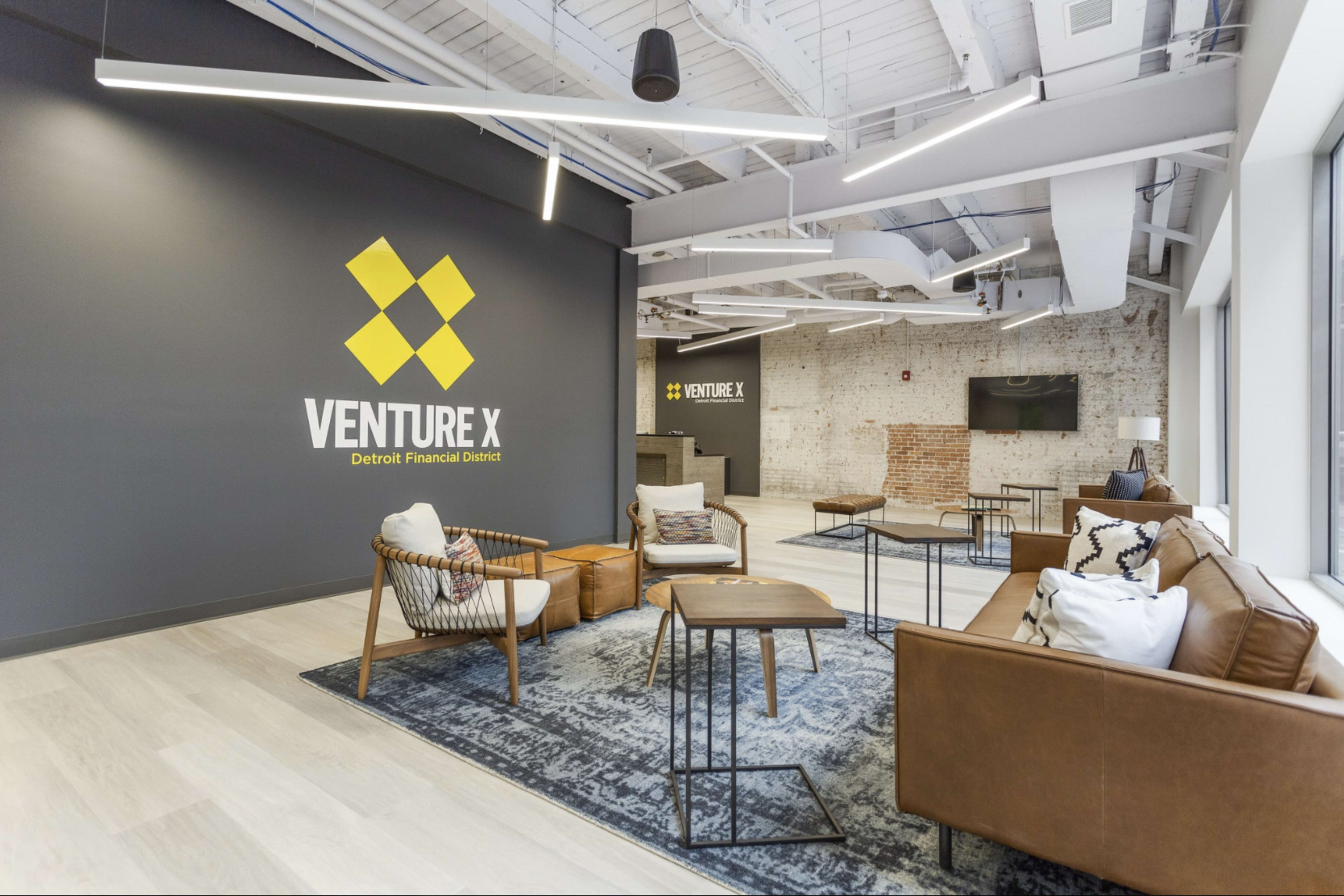 A modern office interior of Venture X, featuring a seating area with chairs and tables, a large wall logo, and exposed brick in the background.