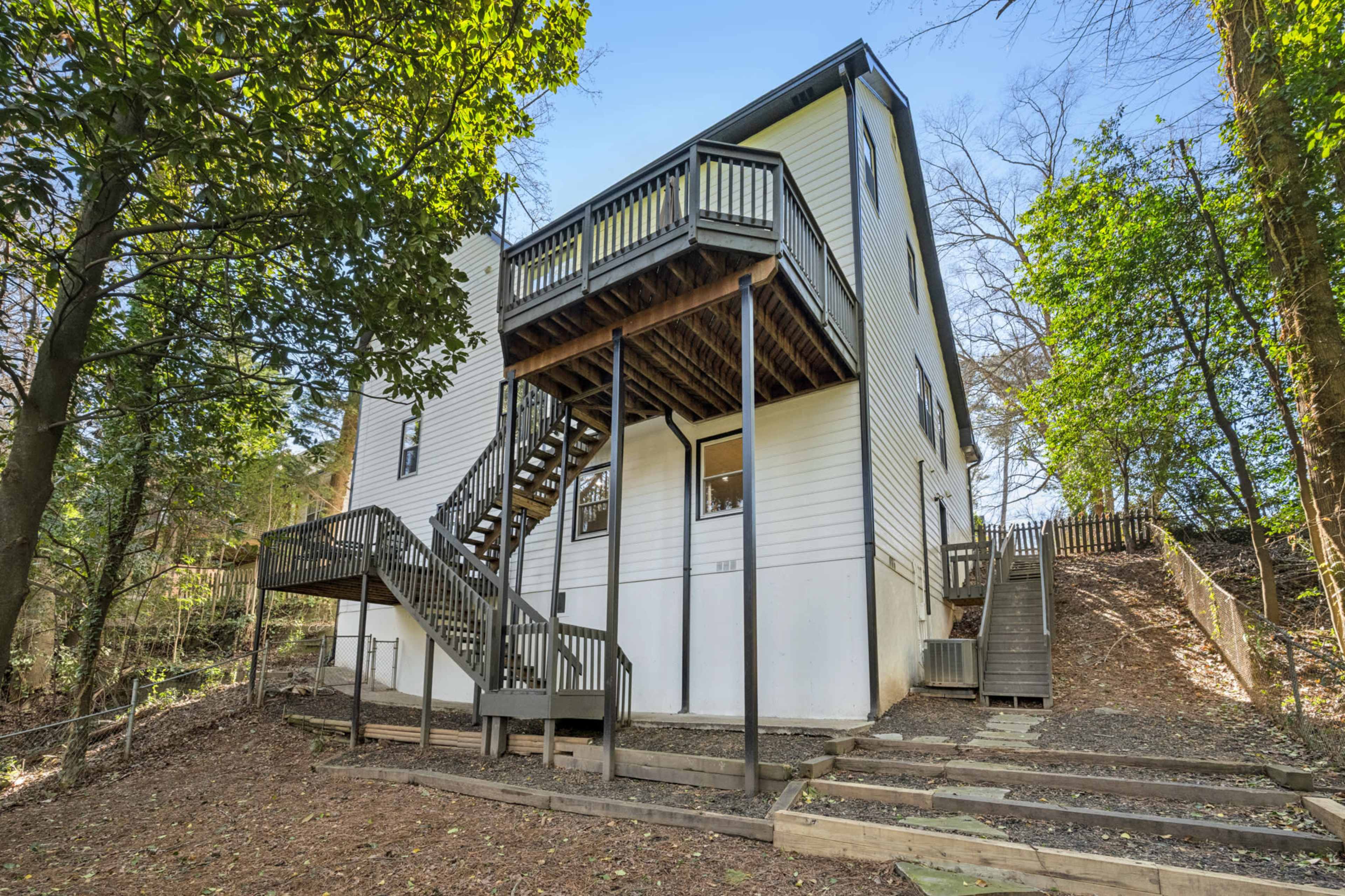 A two-story house with a rear staircase and a wooden deck is surrounded by trees and a sloped yard.