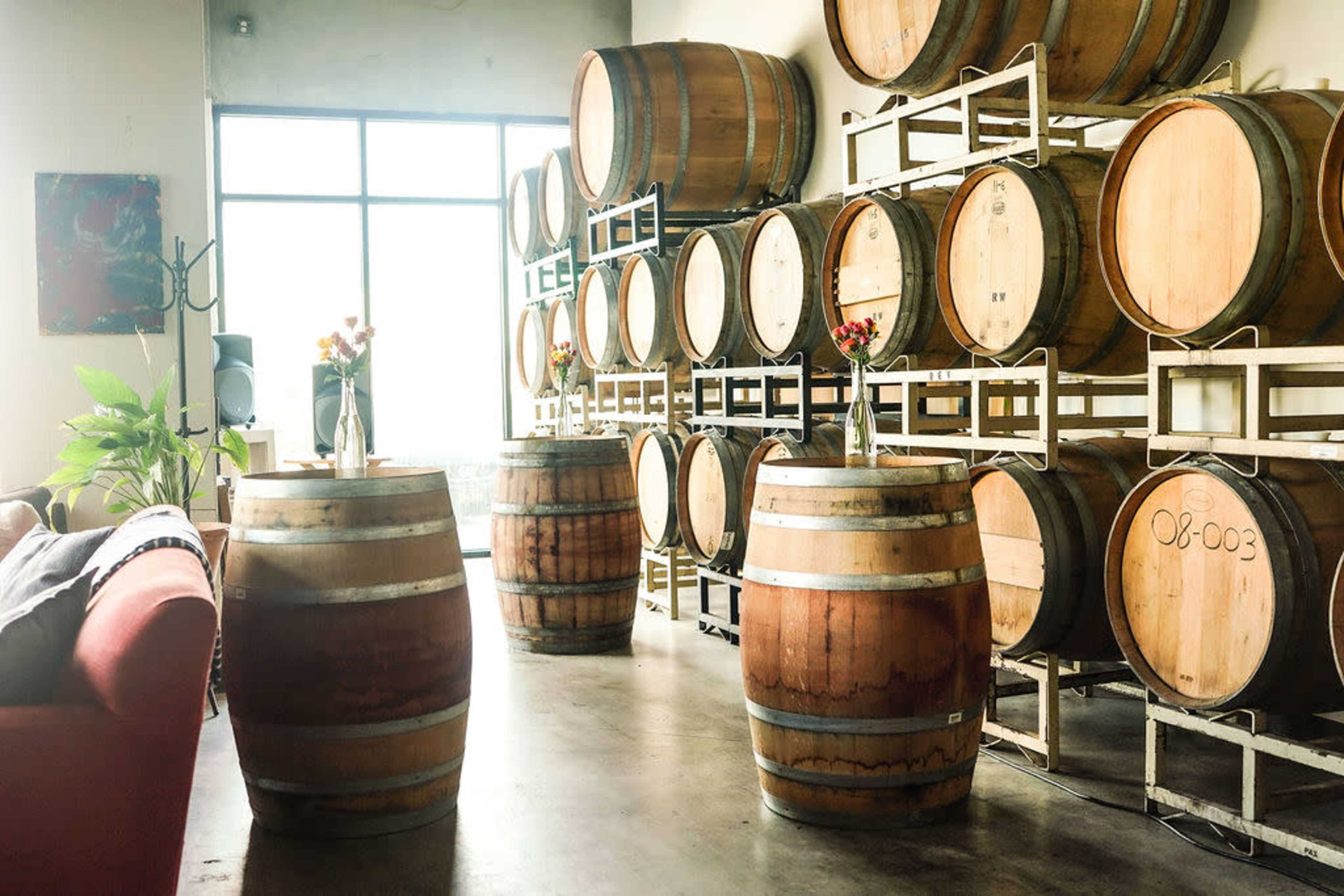 The image shows several wooden barrels arranged against a wall in a modern space, with some barrels serving as tables and decorative vases placed on top.