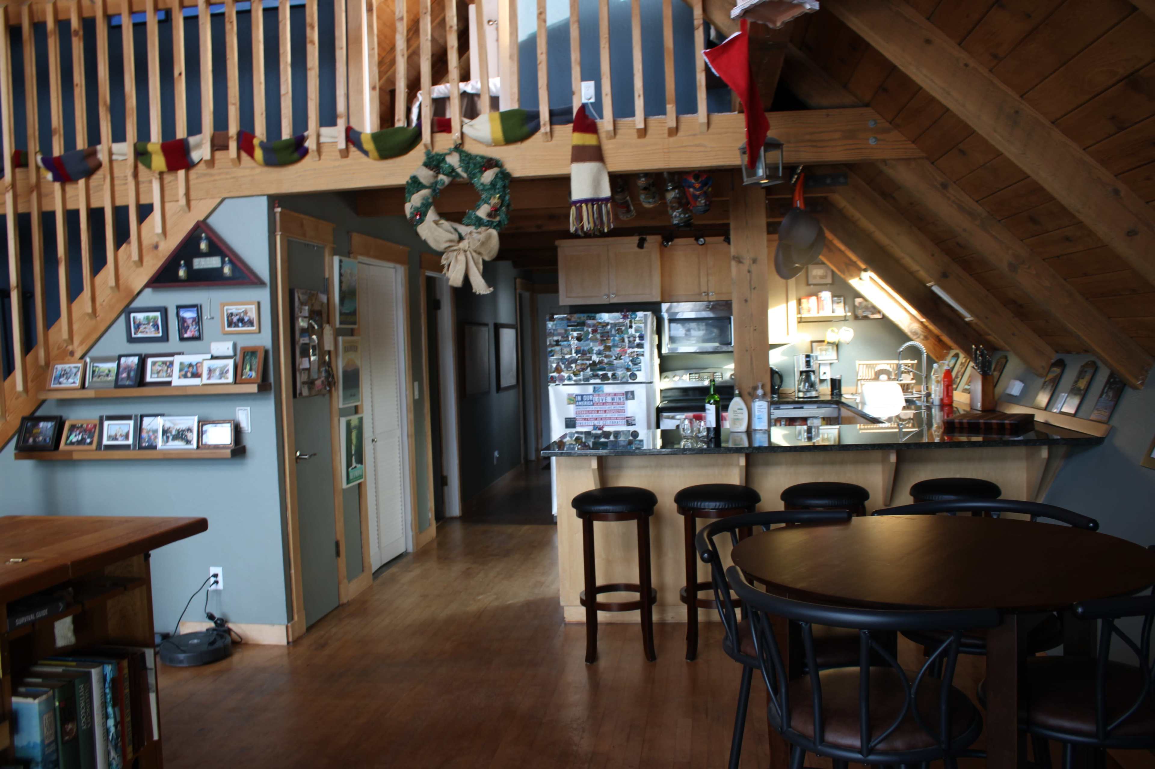 The image shows a wooden interior of a loft-style living space with a kitchen, dining area, and a staircase leading to an upper level.