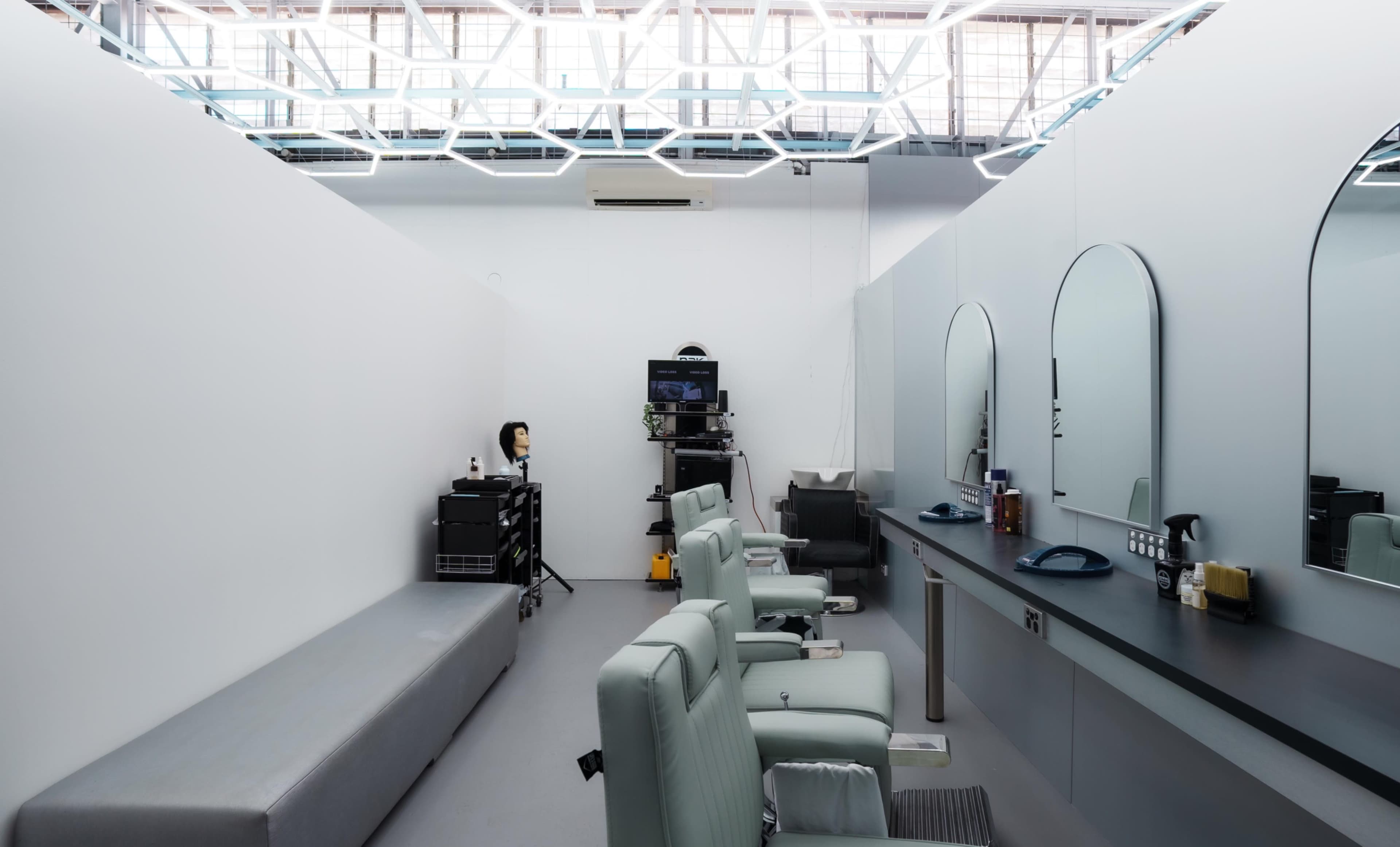 The image shows a minimalist hair salon with several styling chairs, large mirrors, and a wall-mounted television in a well-lit, spacious room.