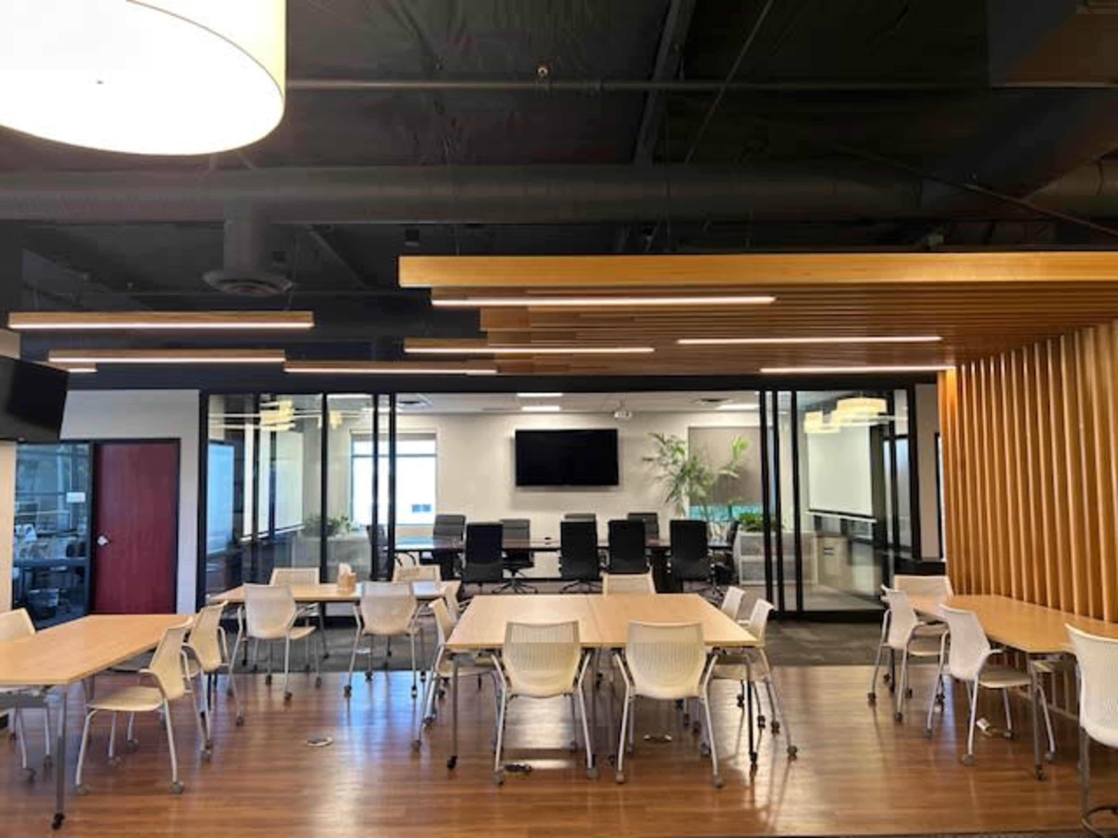 A modern office space featuring multiple tables and chairs, with a conference room visible through glass walls in the background.