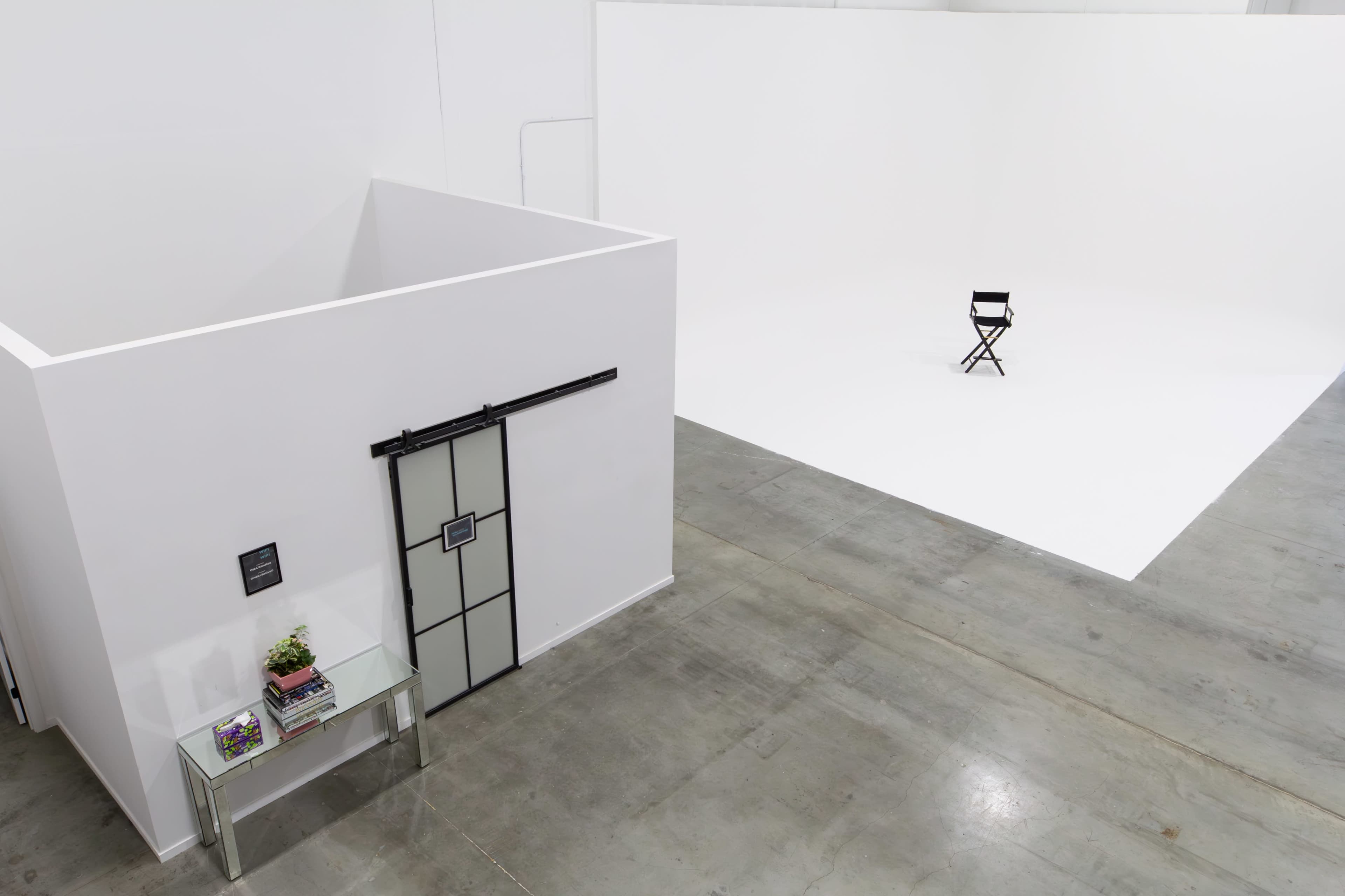 The image shows a simple studio space with a sectioned-off area featuring a sliding door and a small table, along with an empty white backdrop and a single black director's chair in the foreground.