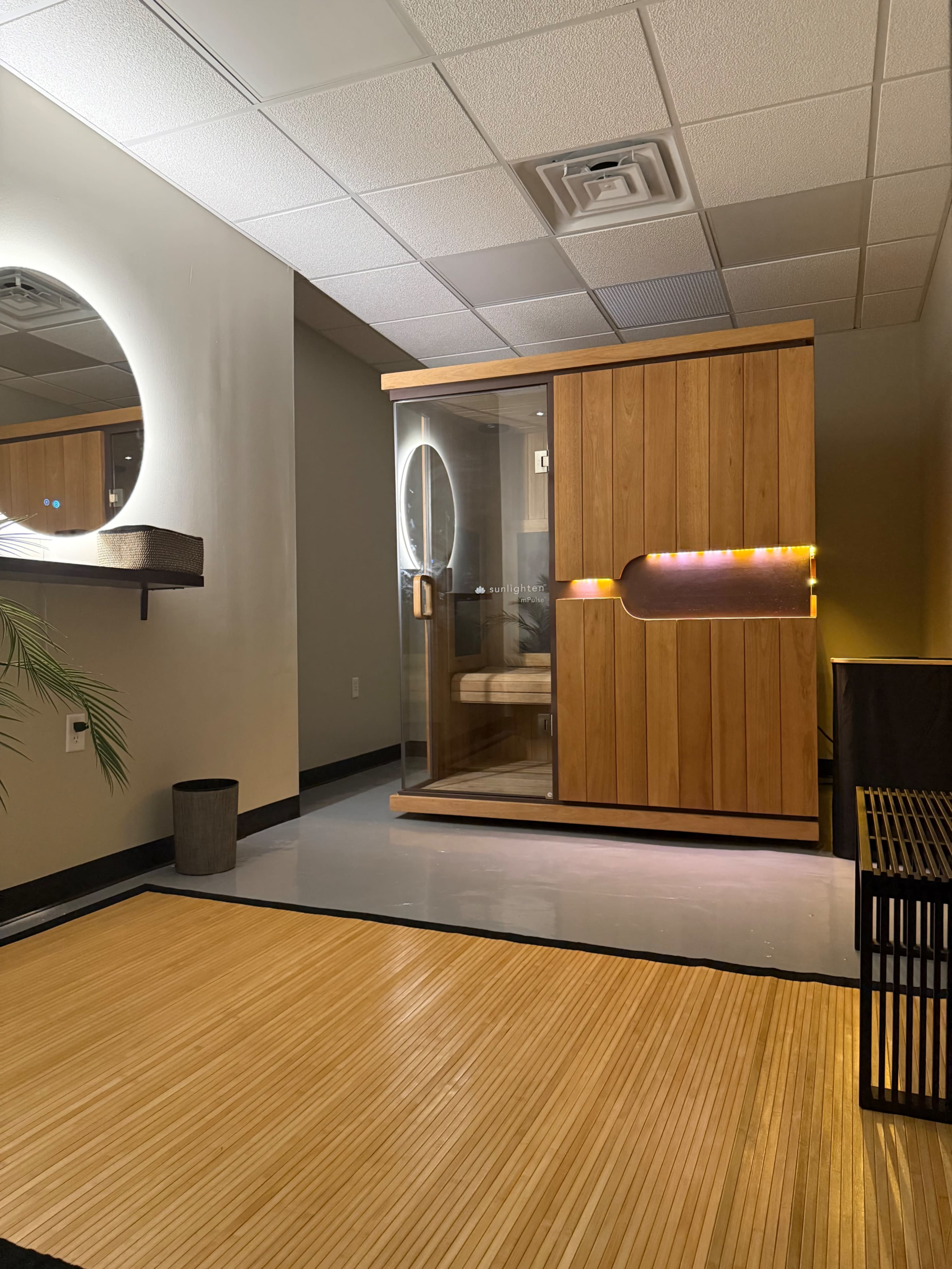 The image shows a modern indoor space featuring a wooden sauna with glass doors, adjacent to a round mirror and a minimalist wooden shelf.