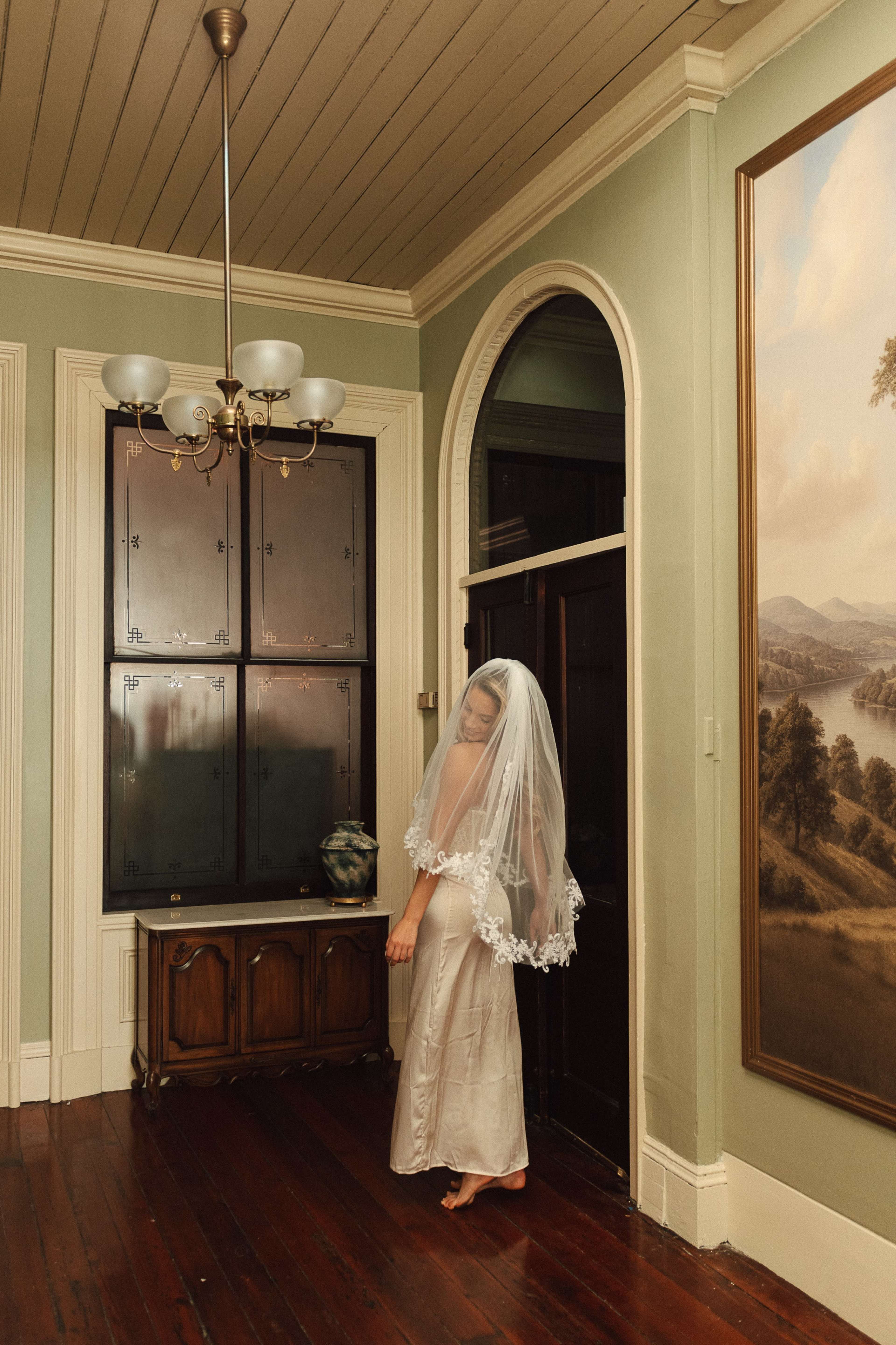 A woman in a wedding dress and veil stands near a dark door in a vintage-style room with wooden floors and ornate decor.