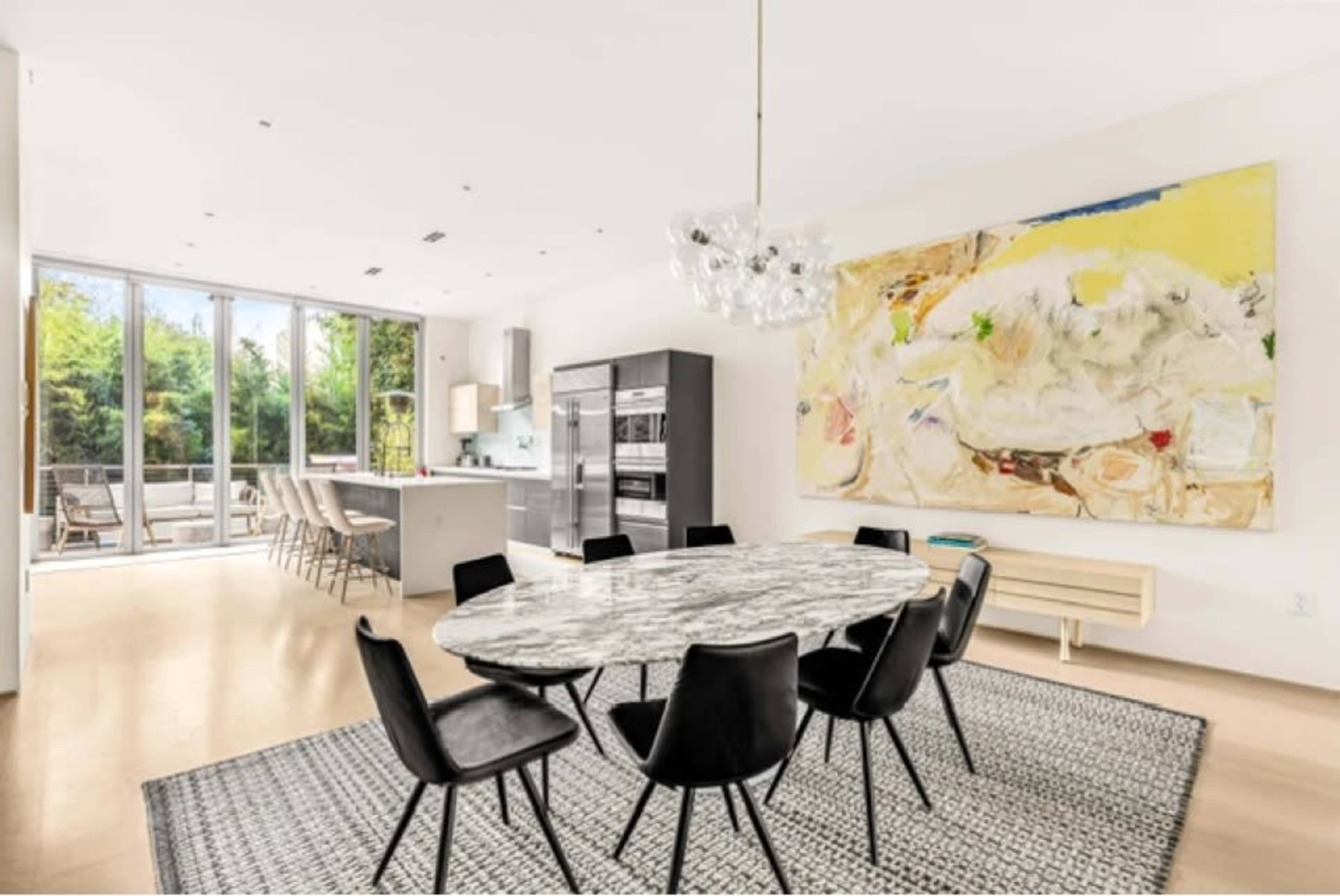 A modern dining area features a round marble table surrounded by black chairs, with large windows and an abstract painting on the wall.