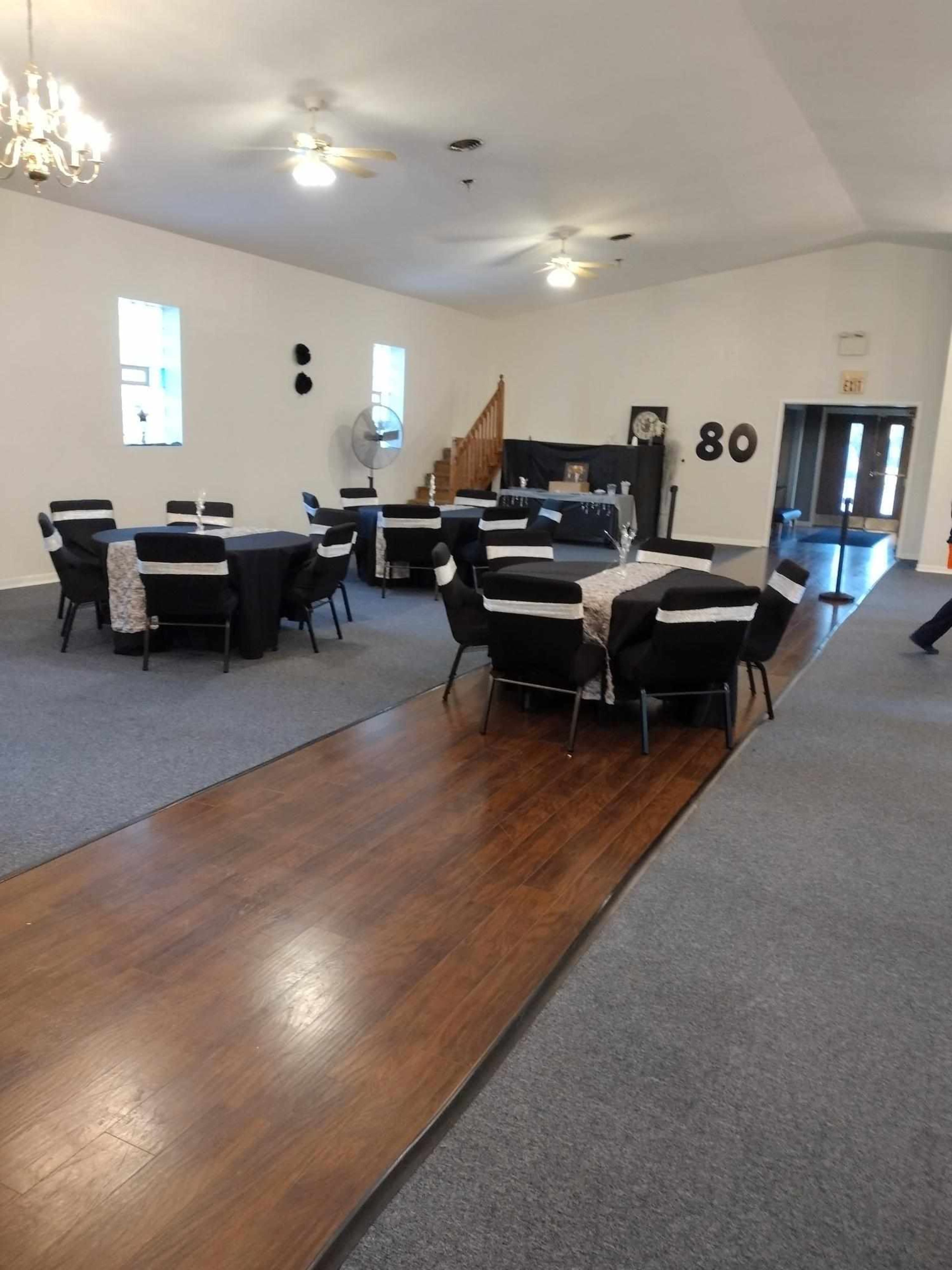 A spacious event hall arranged with tables covered in black and white tablecloths, set against a backdrop of a wooden staircase and decorative wall numbers.