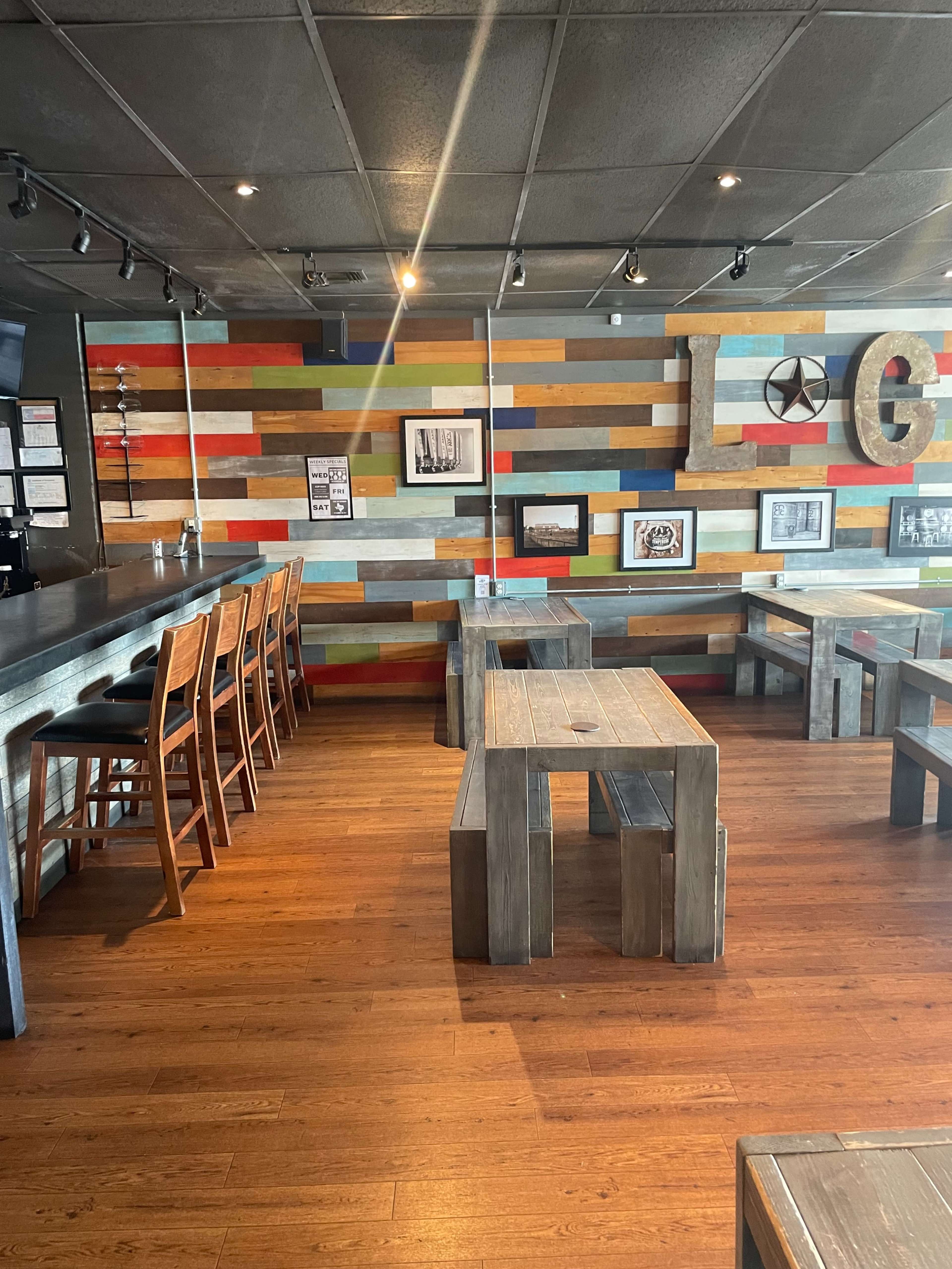 A restaurant interior featuring wooden benches and tables against a colorful, slatted wall, with a bar area on the left.