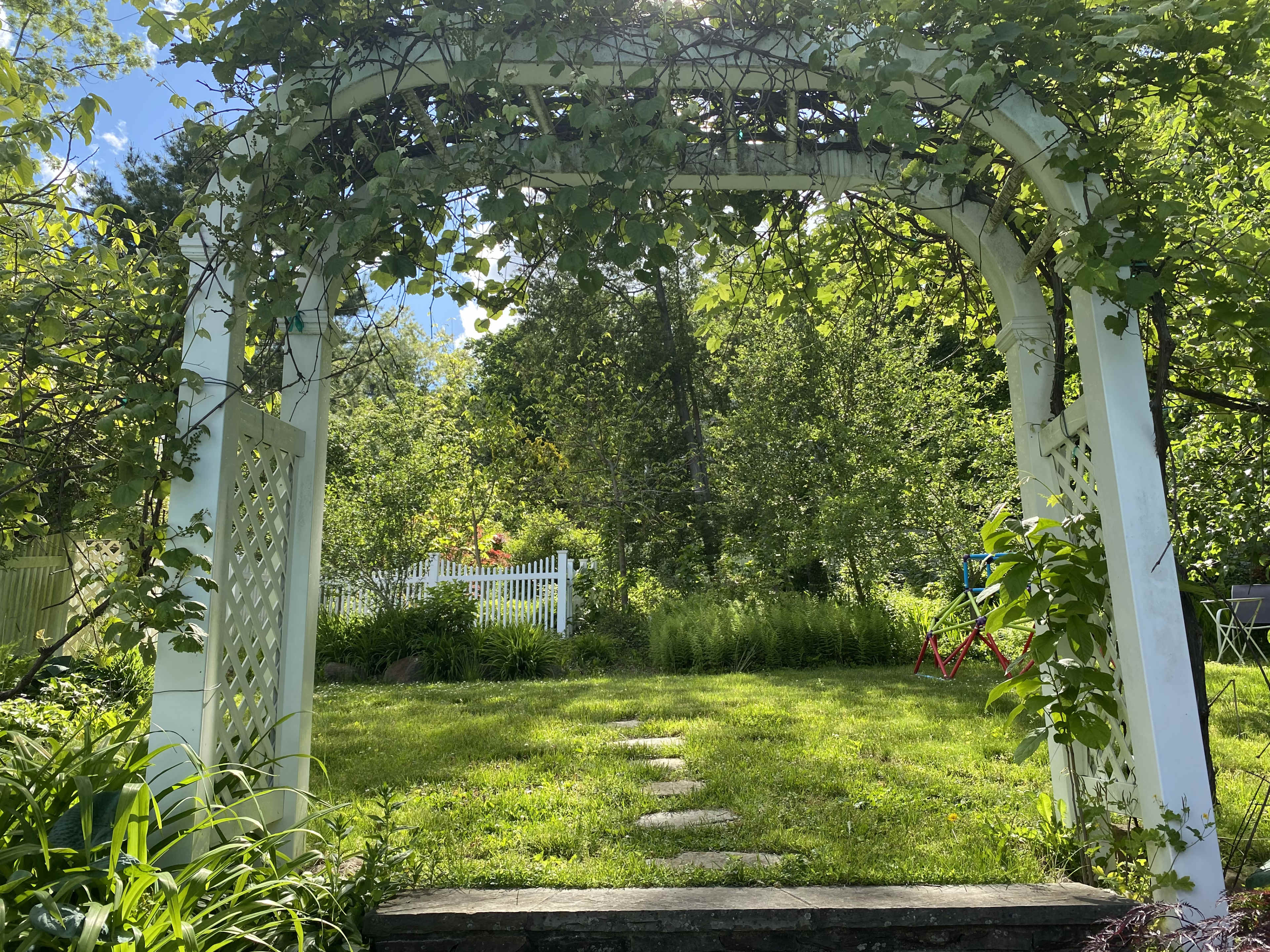 A white wooden arch covered in vines frames a grassy garden with a stone pathway and a white fence in the background.