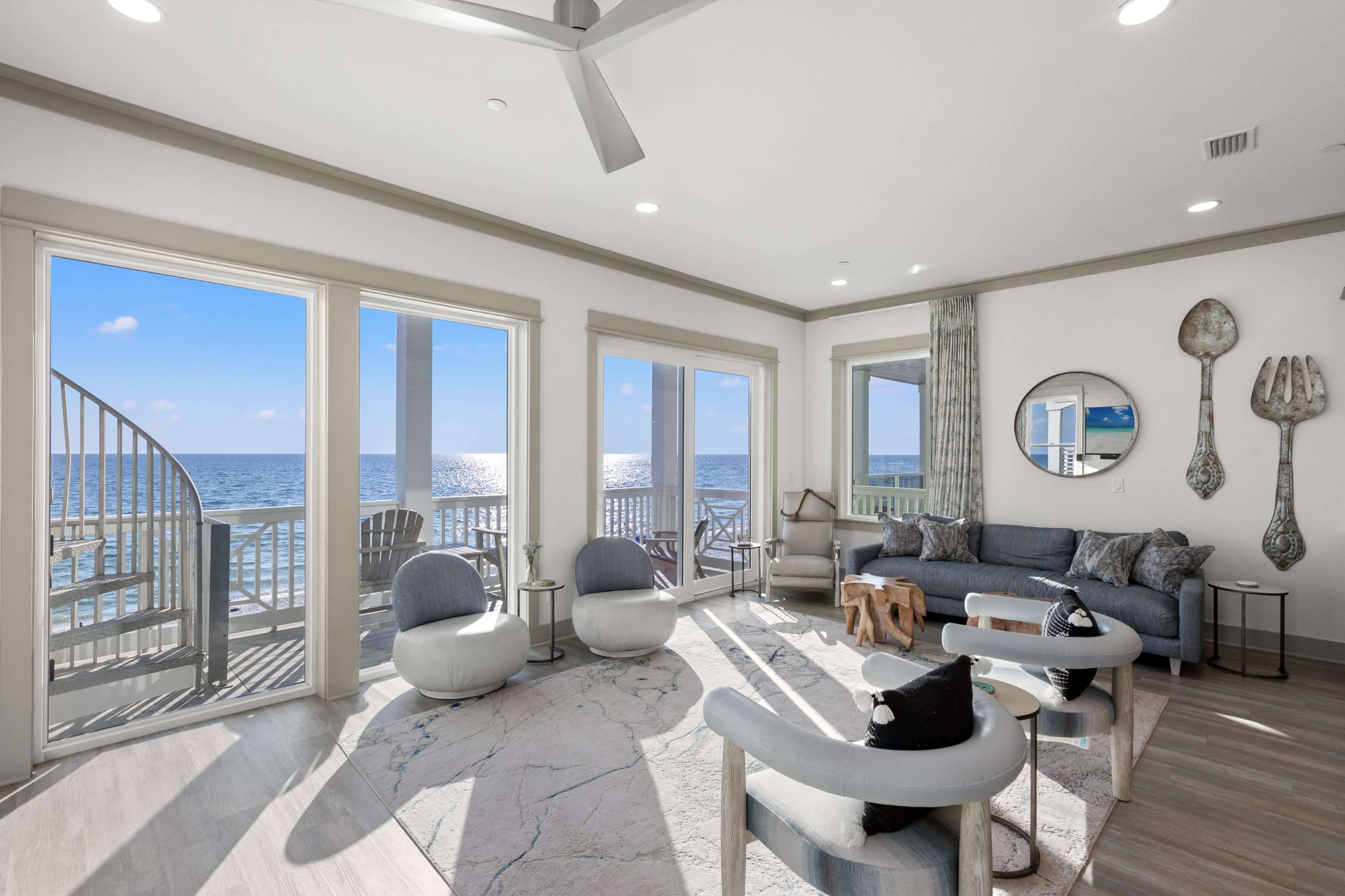 The image shows a modern living room with large windows overlooking the ocean and contemporary furniture, including a sofa and two armchairs.
