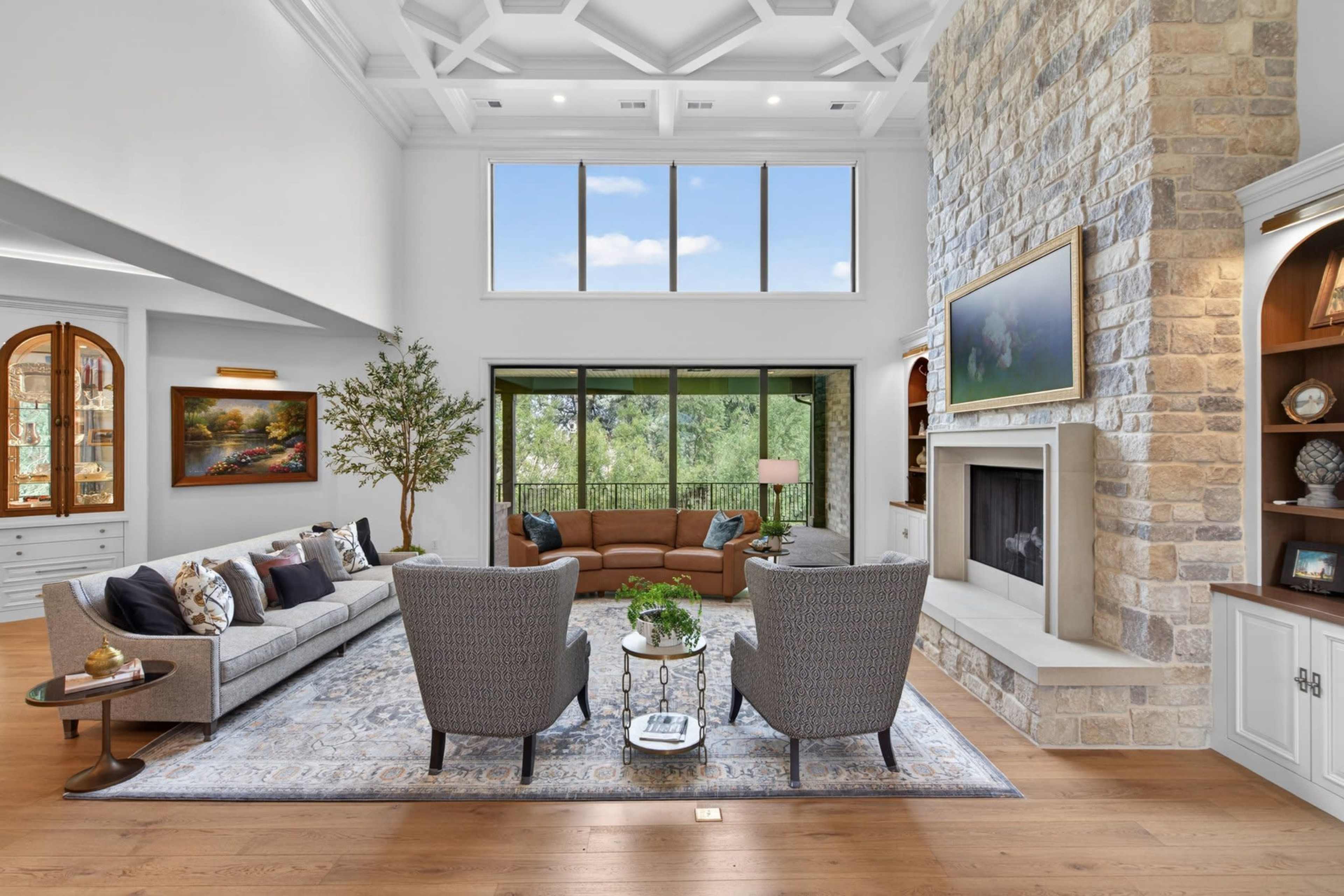 The living room features a high ceiling, large windows overlooking greenery, a stone fireplace, and a comfortable seating arrangement.