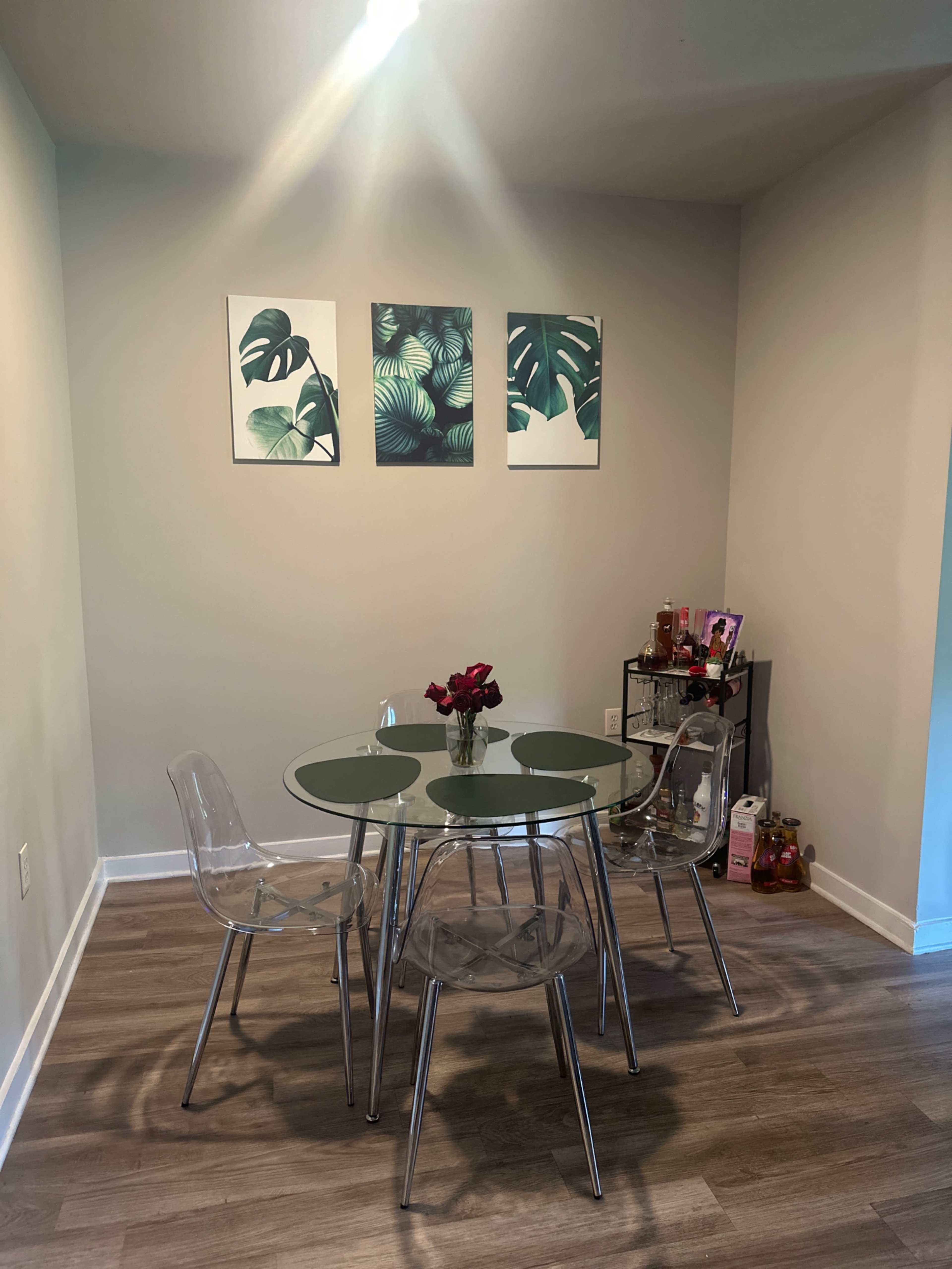 A glass-top dining table surrounded by four clear chairs is set against a wall decorated with three tropical-themed art pieces.