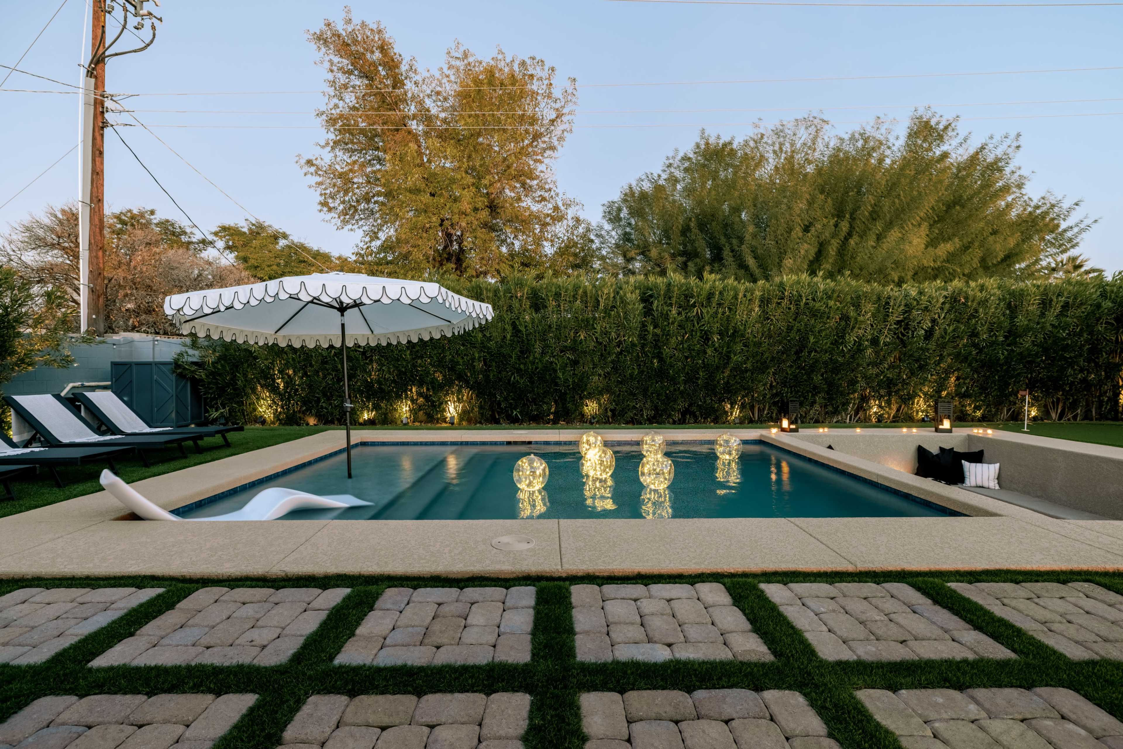 The image shows a modern swimming pool surrounded by lounge chairs, a large umbrella, and illuminated sculptures on the water's surface, with landscaped greenery in the background.
