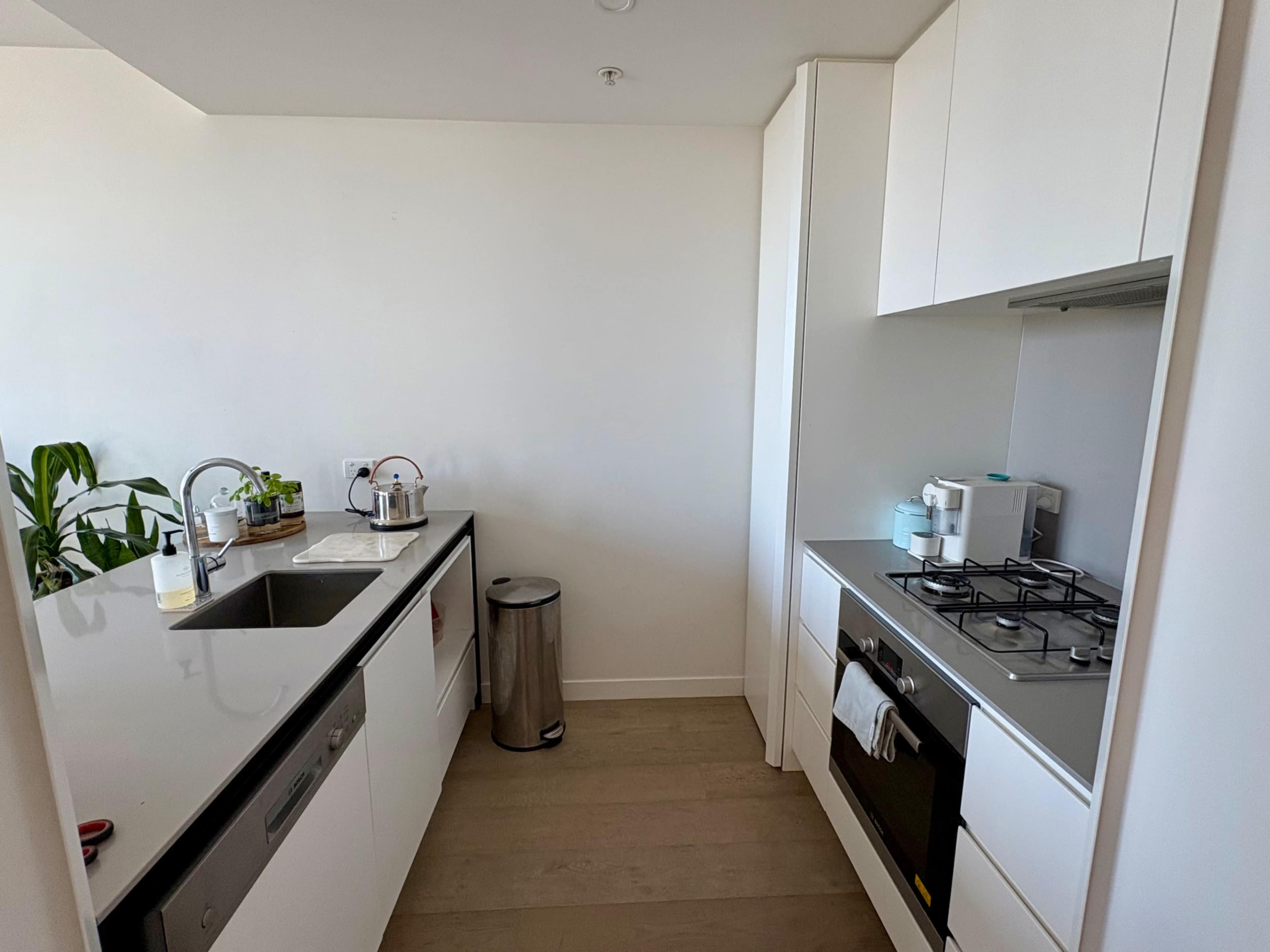 The image shows a modern kitchen with white cabinetry, a stainless steel sink, a gas stove, and a trash can, featuring minimal decor.