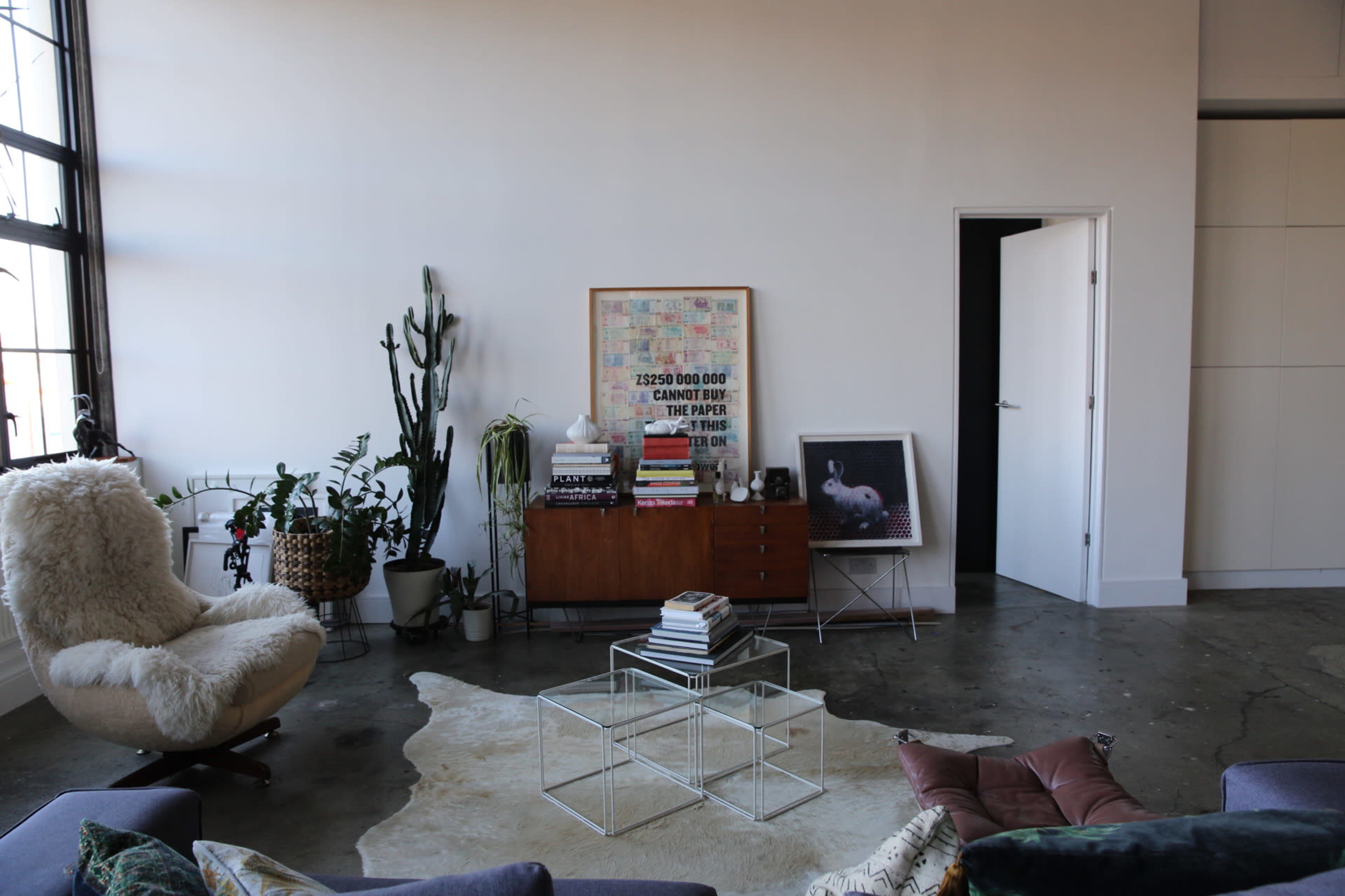 The image features a modern living room with a large window, a cozy chair, a wooden cabinet filled with books, and a decorative rug on a concrete floor.