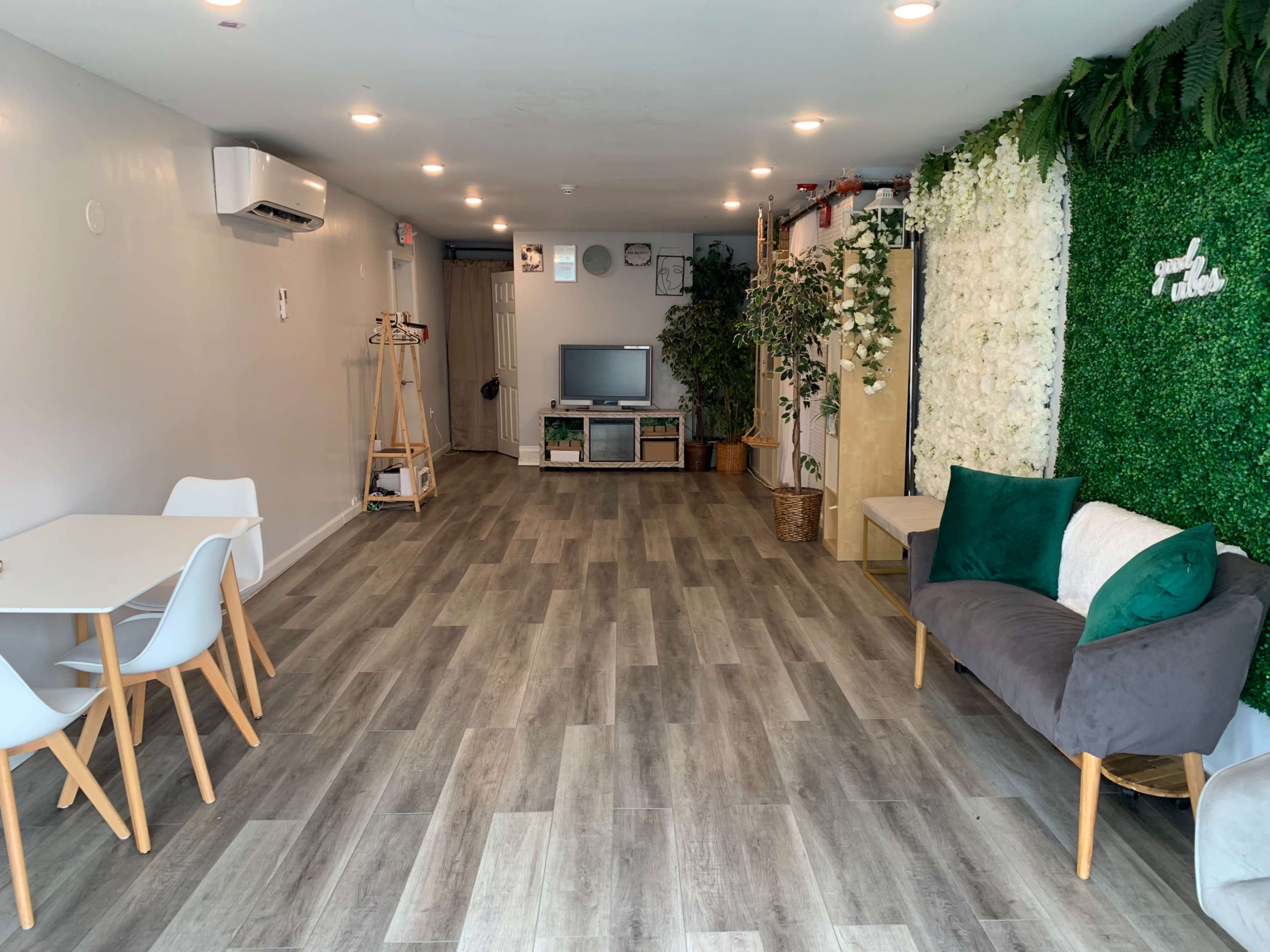 A modern, minimalist interior with a light wood floor, a small dining area with white chairs, a green wall of plants, and a seating area featuring a gray couch and decorative pillows.