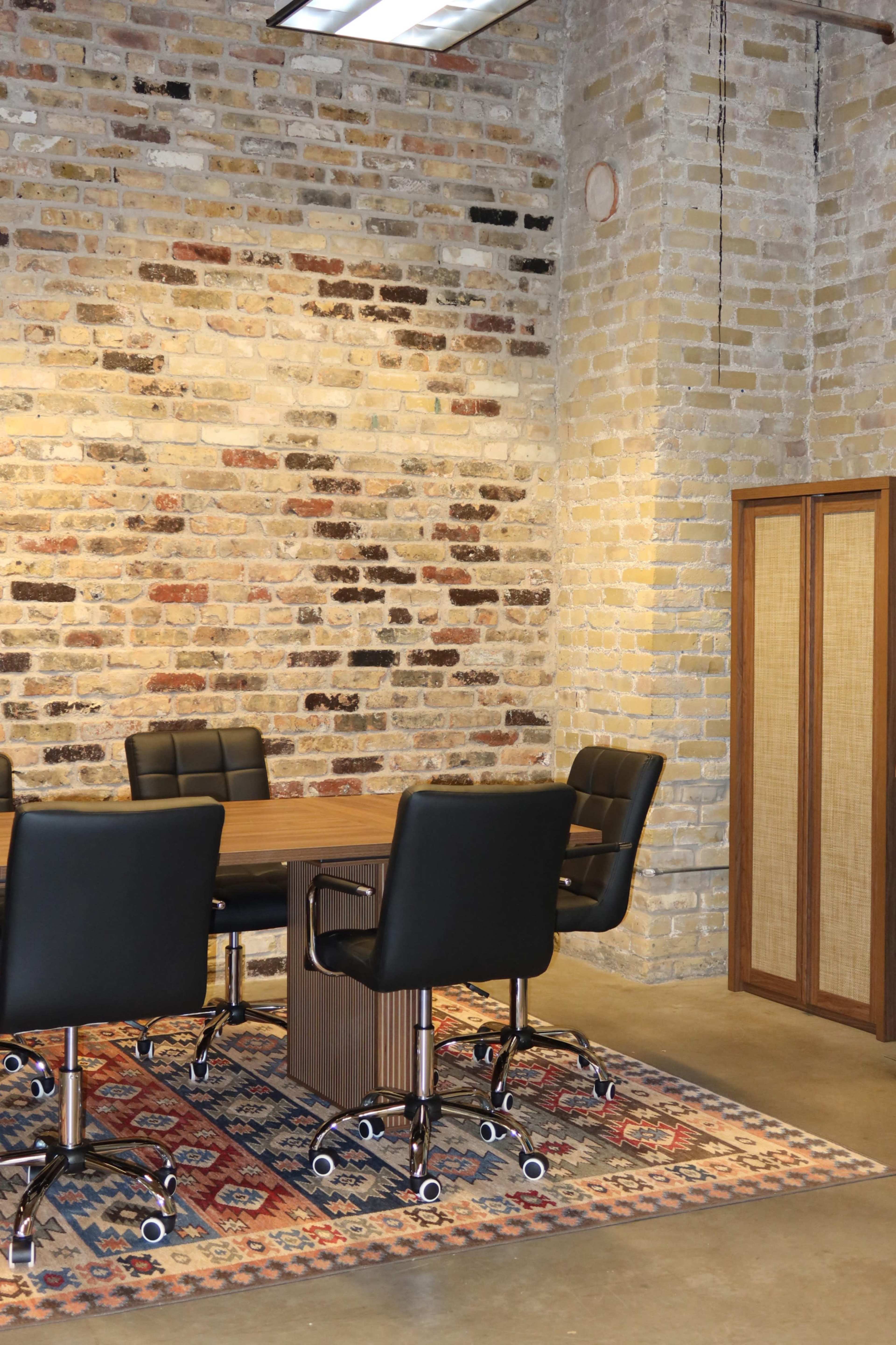 A conference room features a wooden table surrounded by black leather chairs, set against a backdrop of a textured brick wall and a decorative rug.