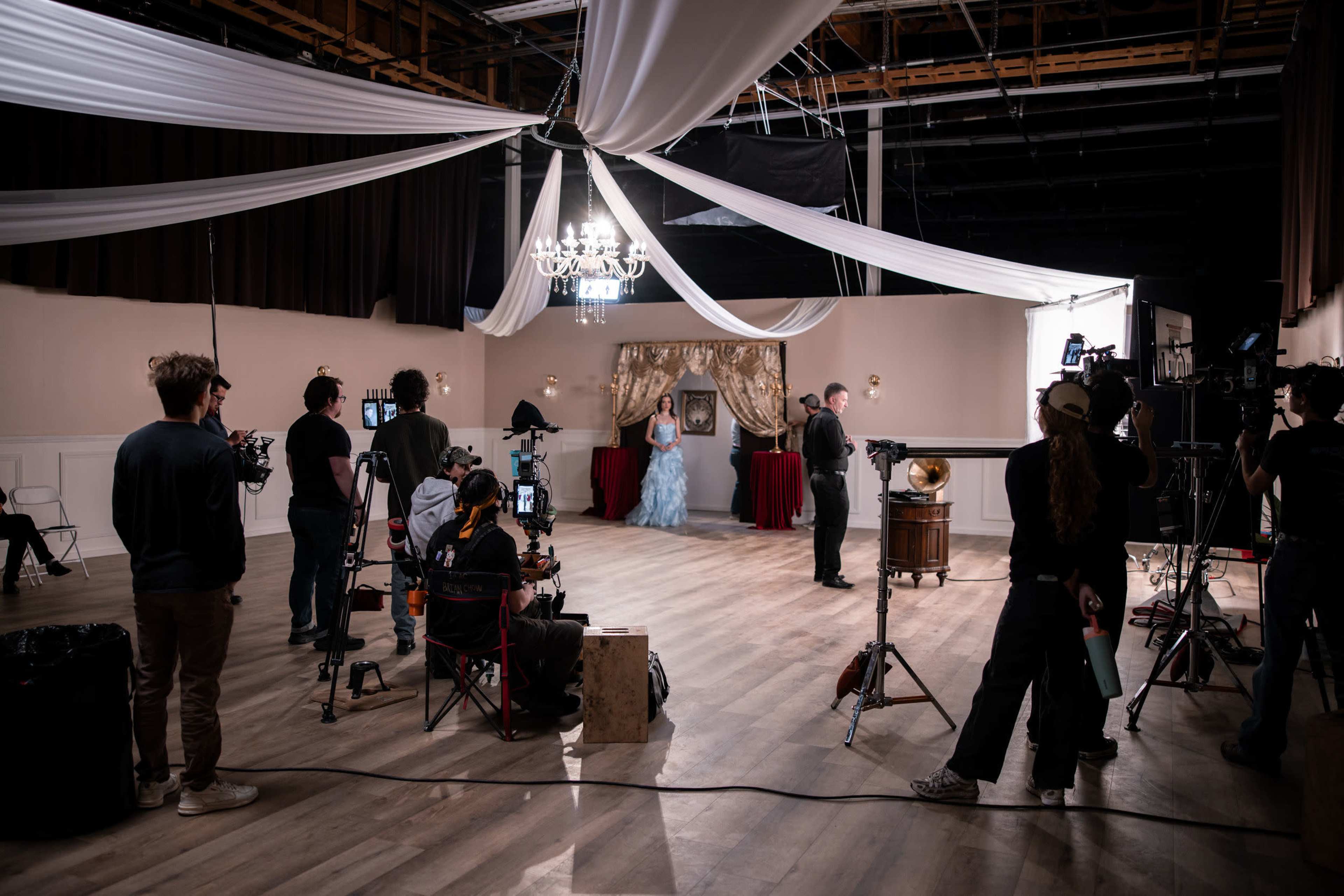 A film crew is set up in a studio, with multiple cameras and lights focused on a woman in a blue dress standing in front of a backdrop.