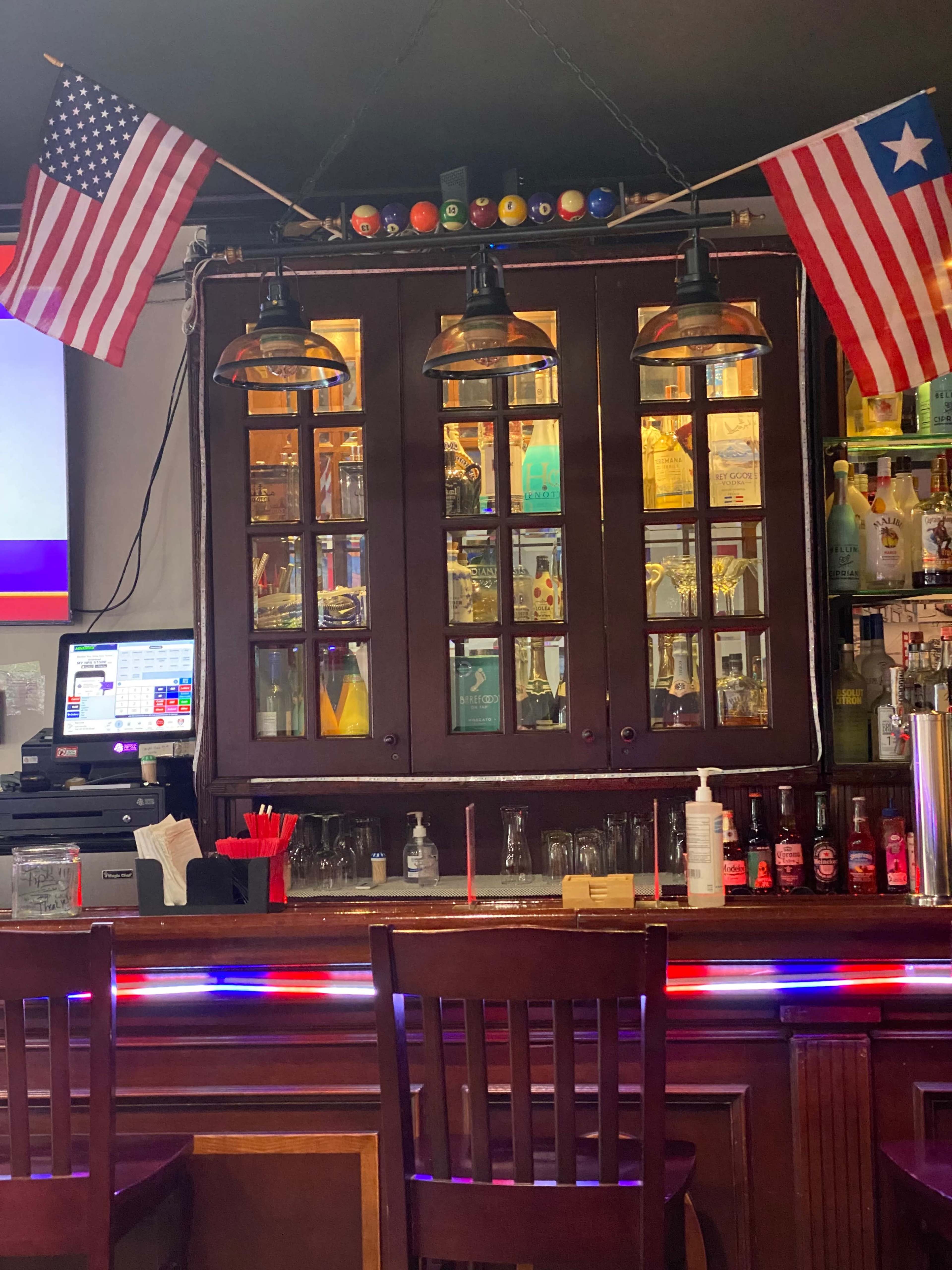 A bar area with a wooden counter, illuminated by hanging lamps, and decorated with American and Texan flags above a glass display cabinet filled with bottles.
