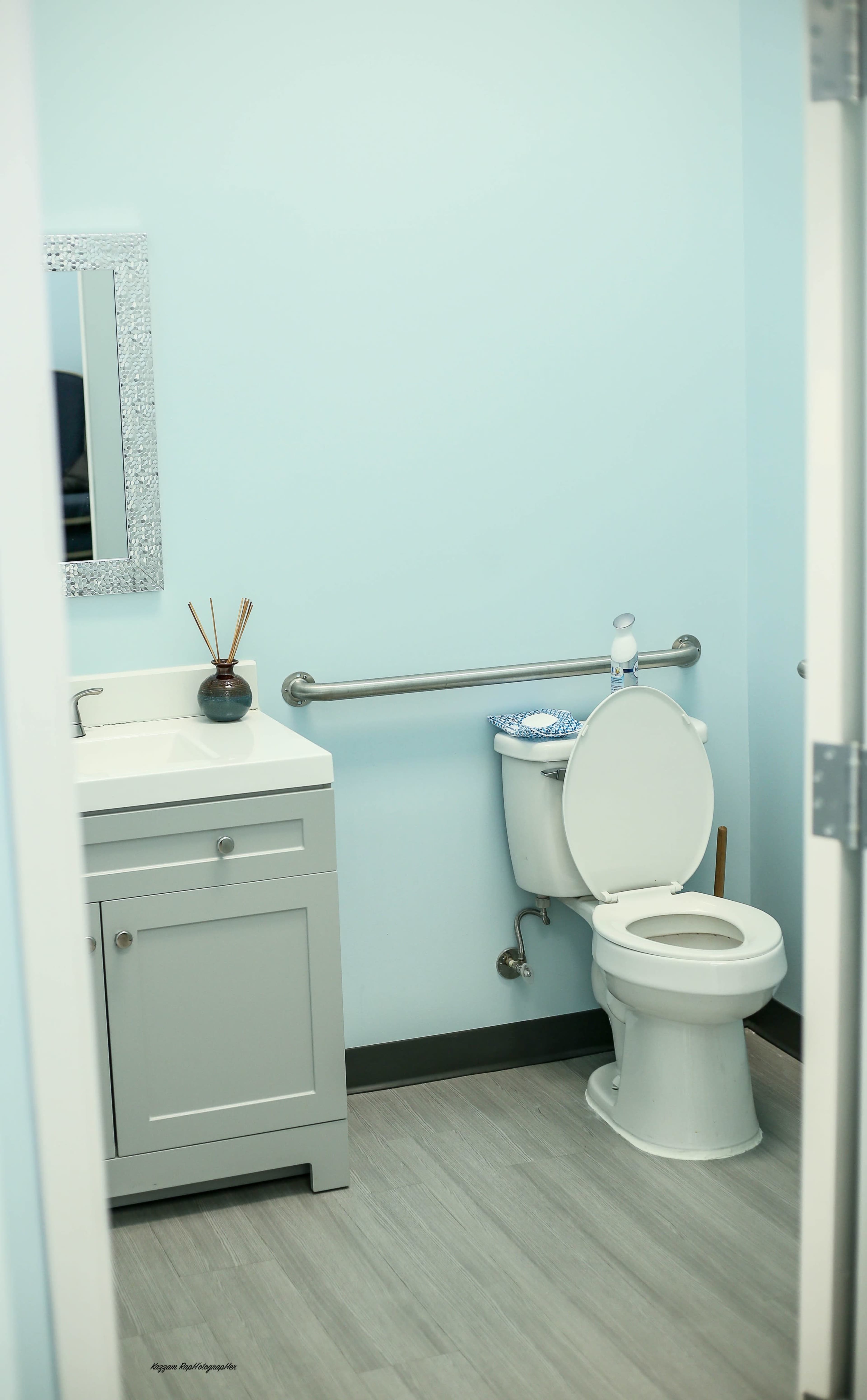 The image shows a small bathroom featuring a toilet, a sink with a cabinet, and a mirror above the sink, all set against light blue walls.