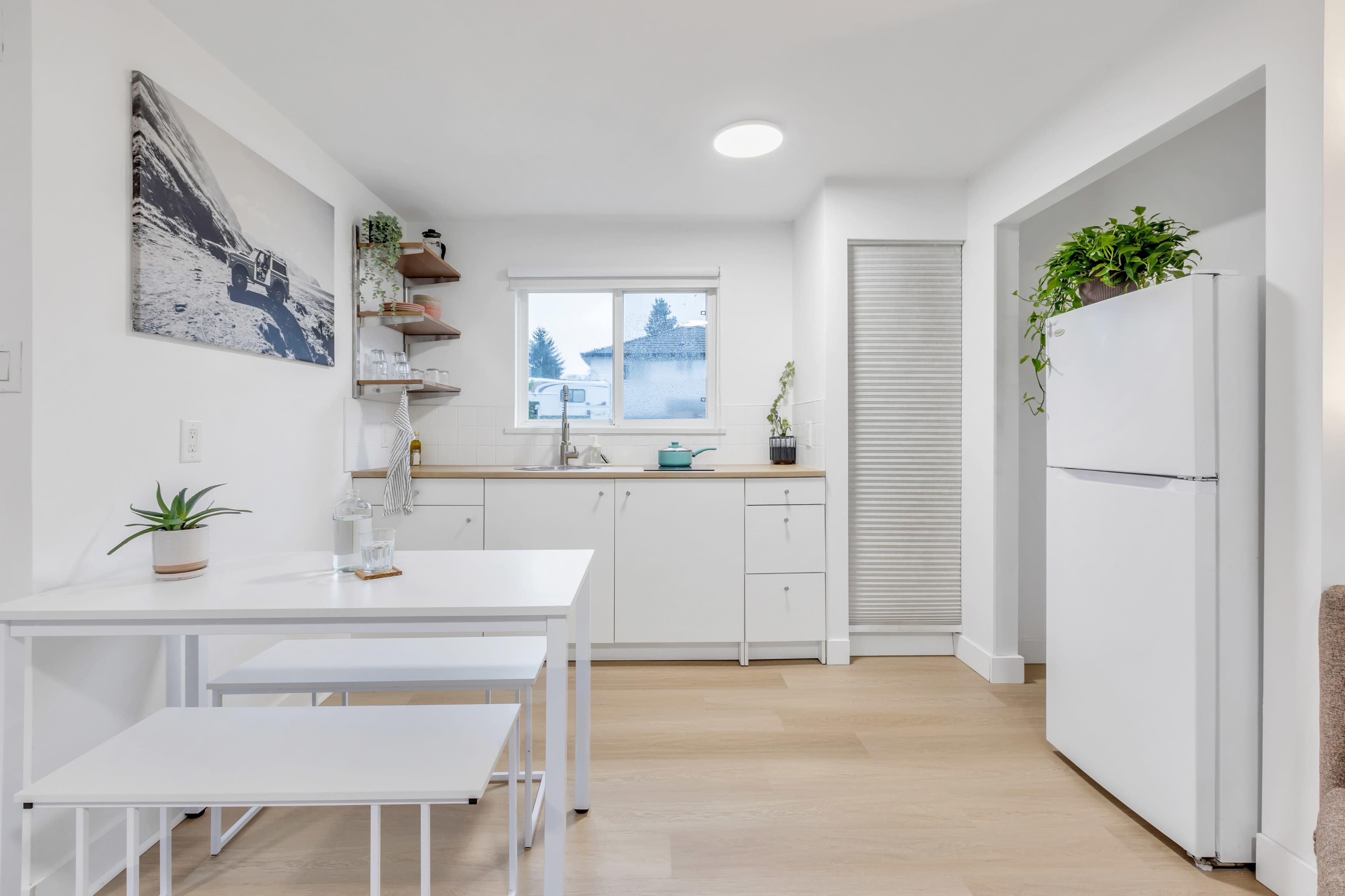A modern kitchen features white cabinetry, a small dining table, and a refrigerator, with a window providing natural light.
