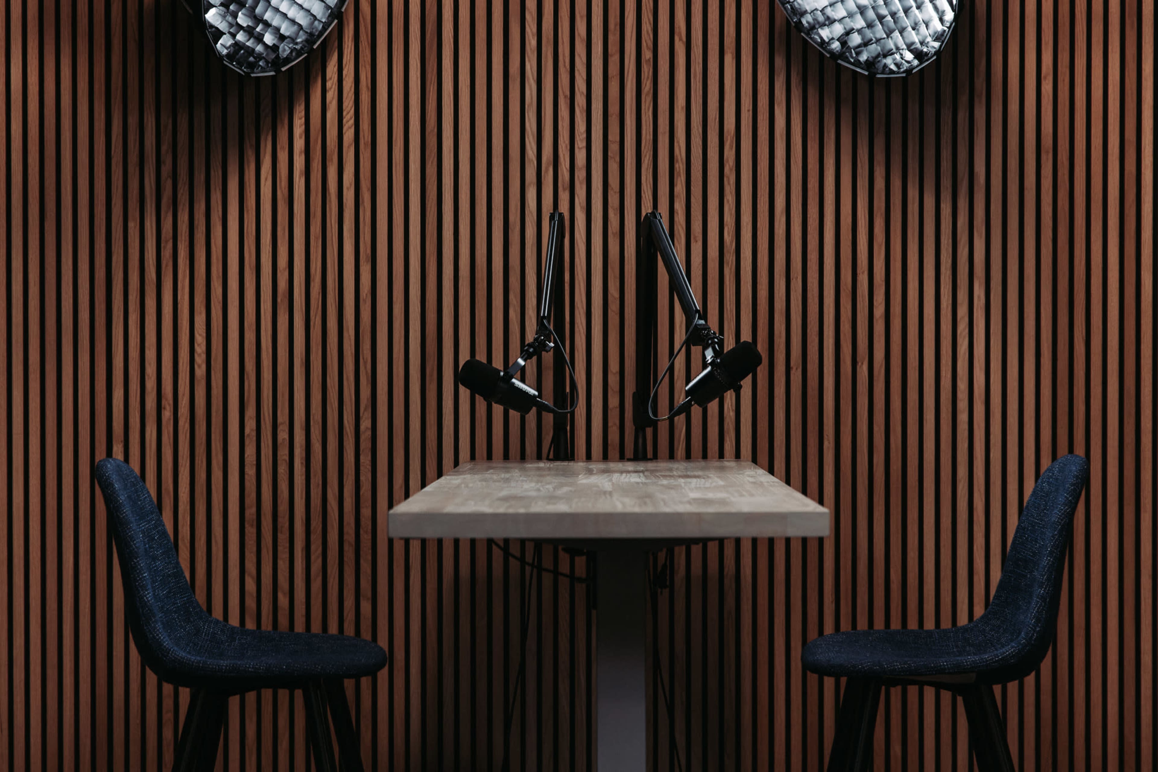 A wooden table with two chairs and microphone setups is positioned against a slatted wooden wall.