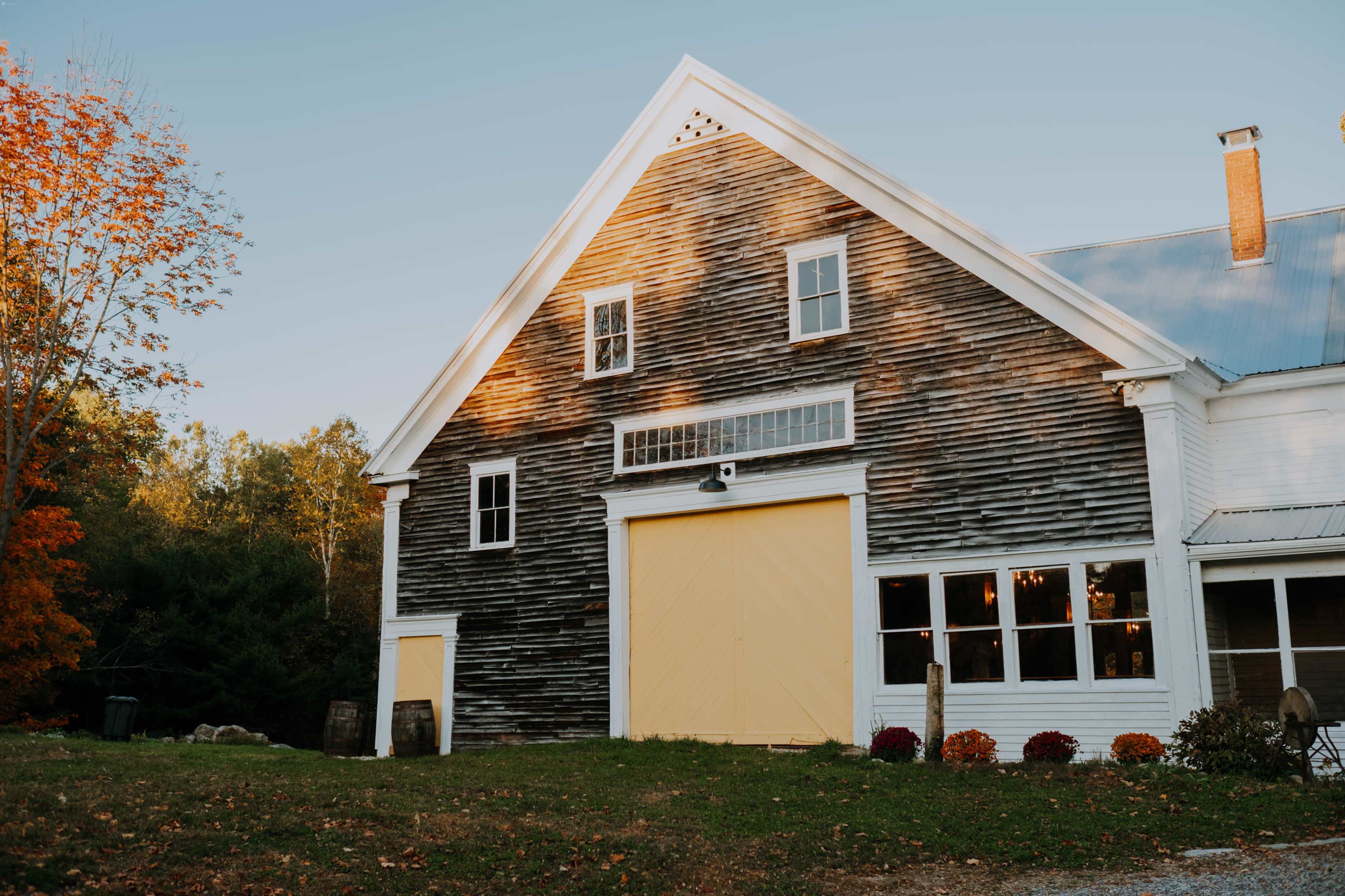 A rustic wooden barn with a yellow door, set against a backdrop of trees and autumn foliage.