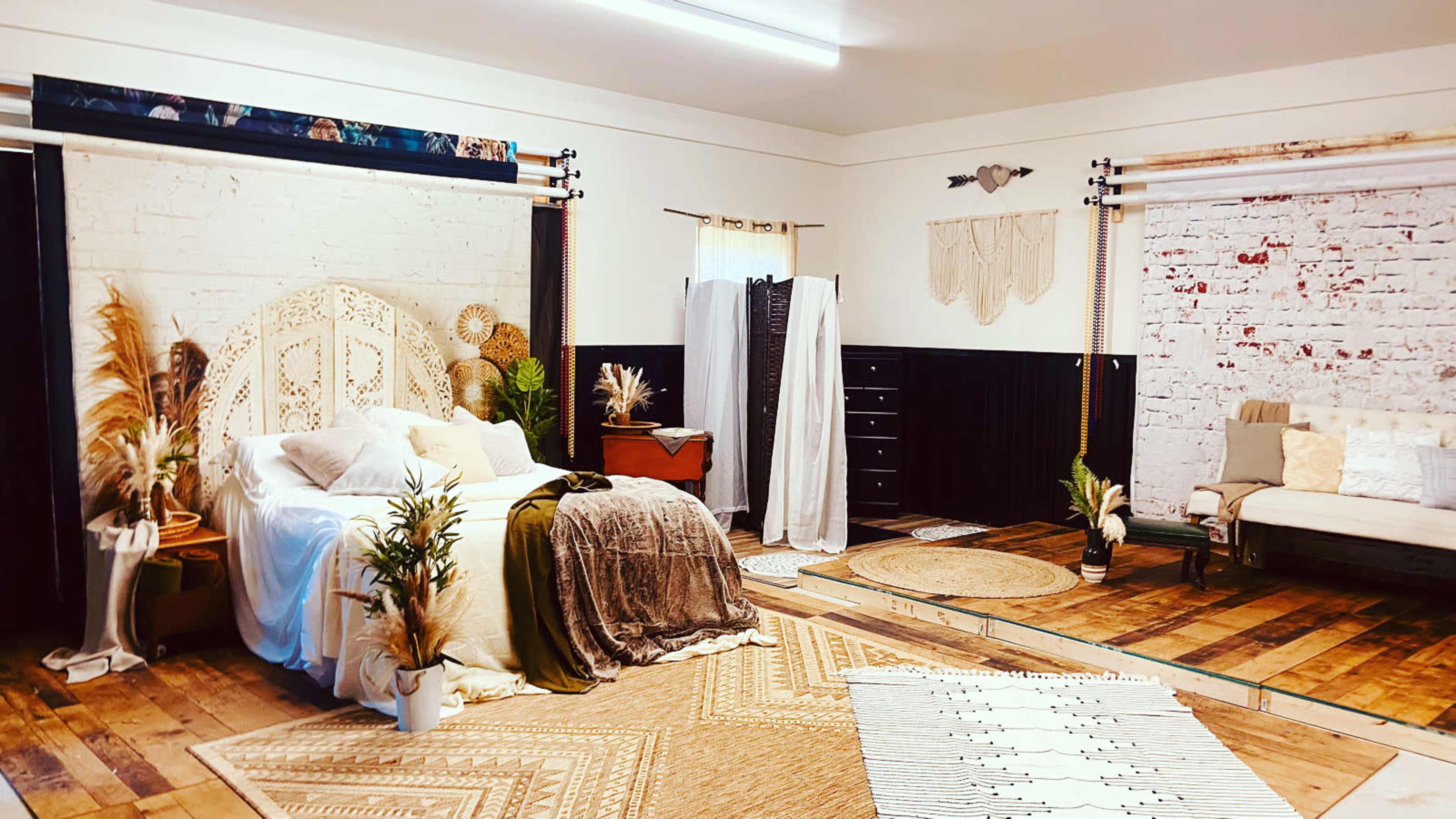 A styled interior room featuring a bed with woven headboard, various decorative plants, and distinct seating areas separated by patterned rugs and backdrops.