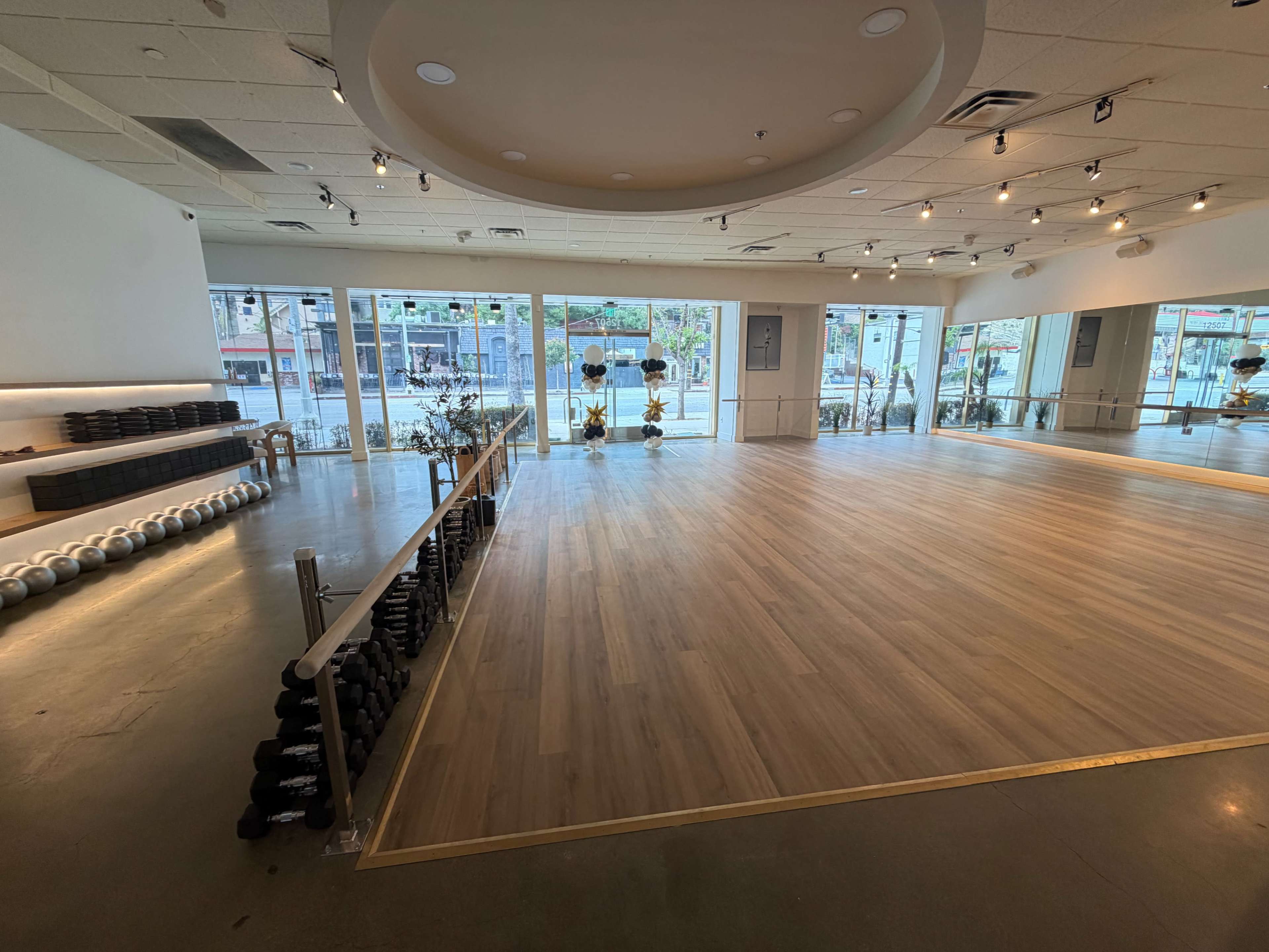 The image shows a spacious fitness studio with wooden flooring, large windows, and neatly arranged dumbbells along the walls.