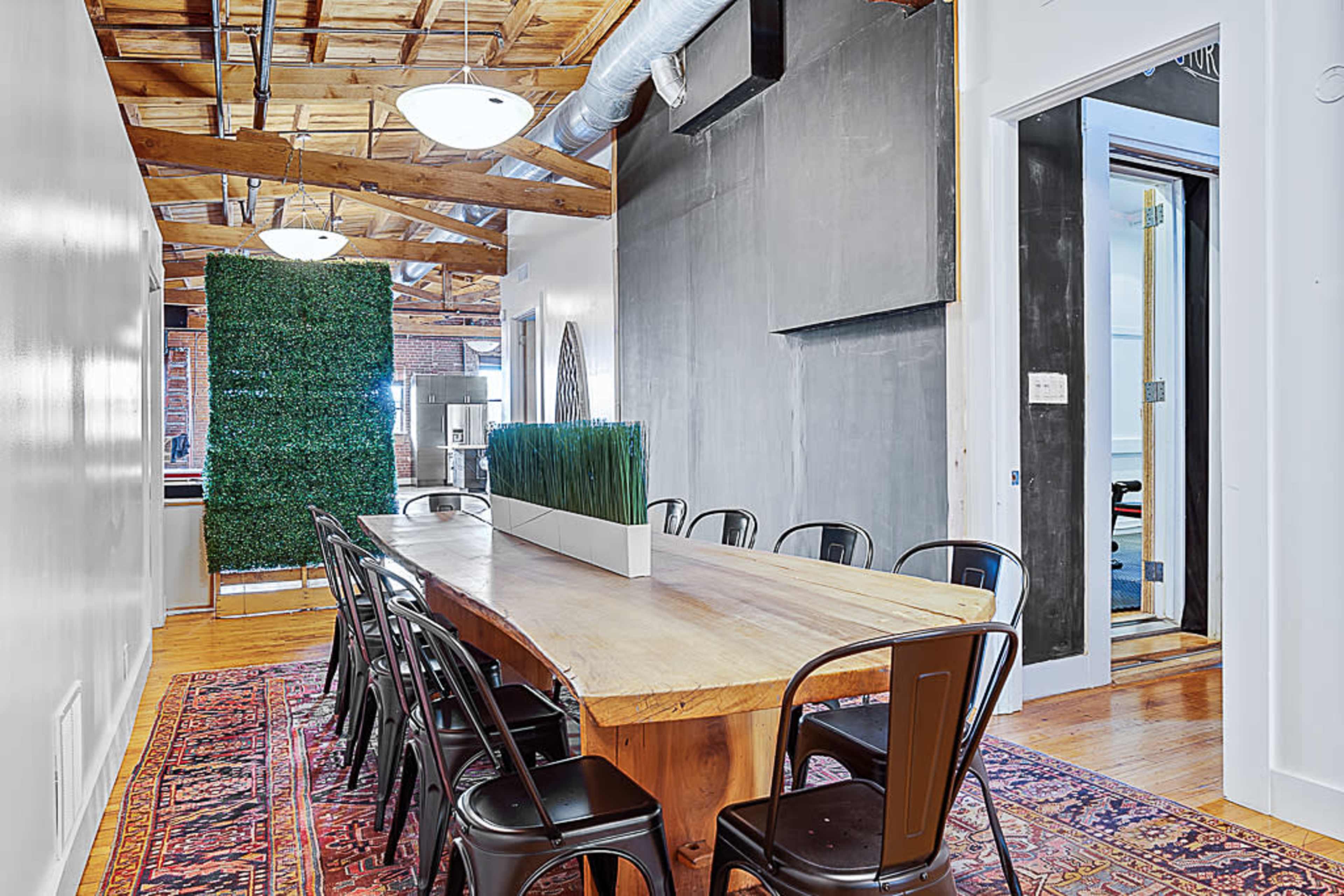 A dining area featuring a large wooden table with black metal chairs, a green plant installation, and a chalkboard wall.