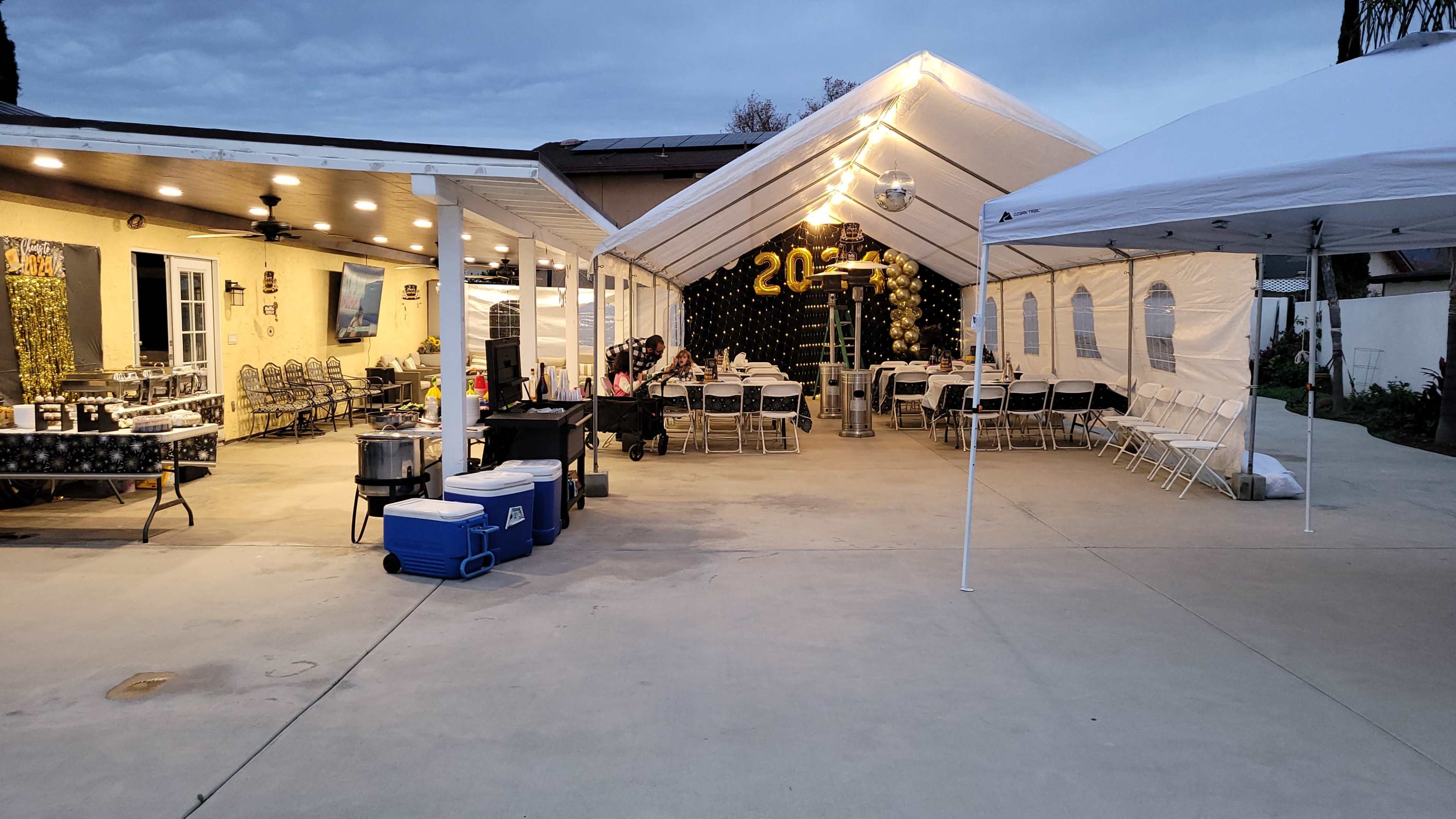 A covered outdoor area with tables and chairs is set up for a gathering, adjacent to a house featuring a food preparation area and decorations.