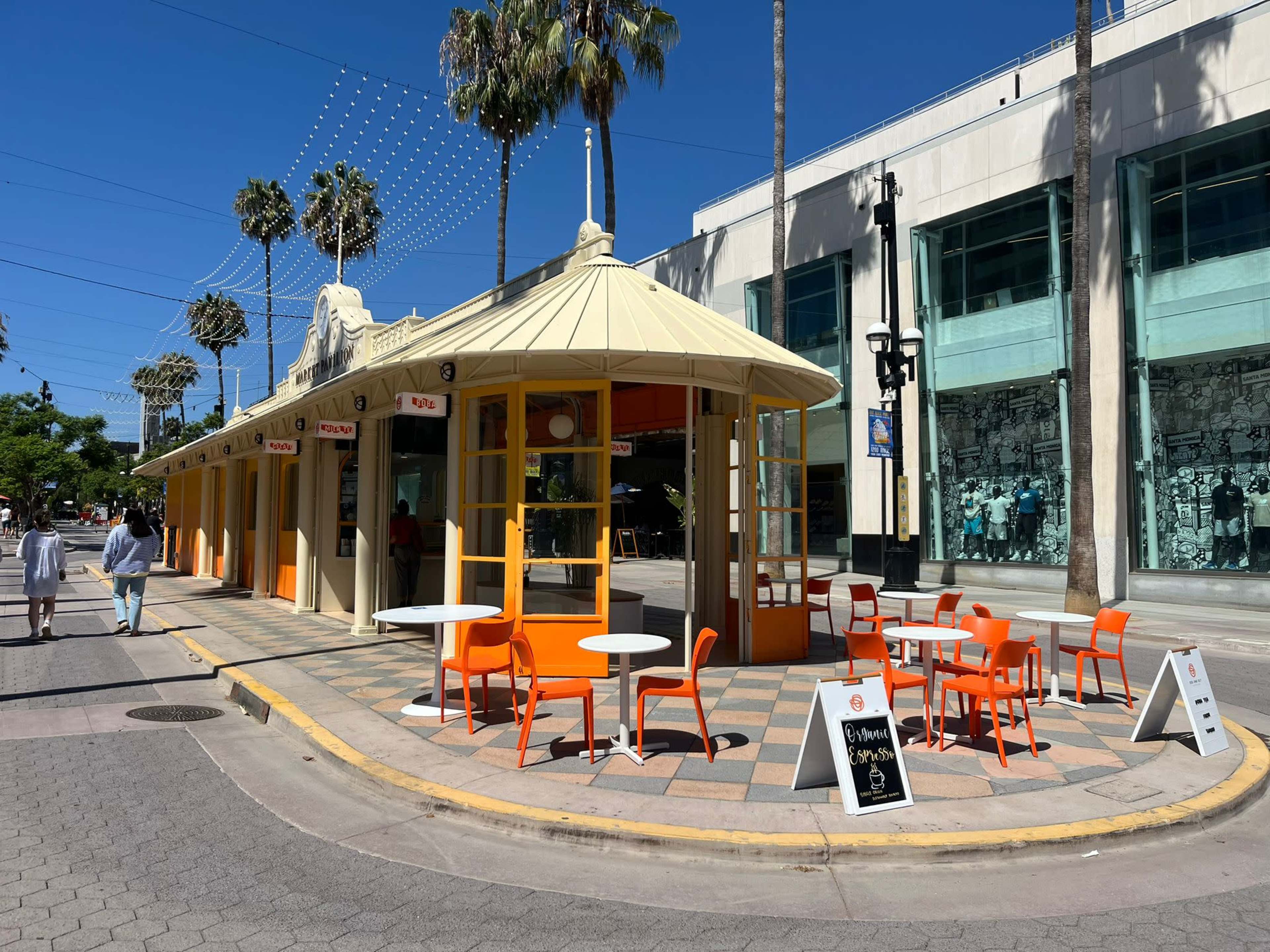 A colorful outdoor café with orange and white furniture is situated on a corner, surrounded by palm trees and modern buildings.