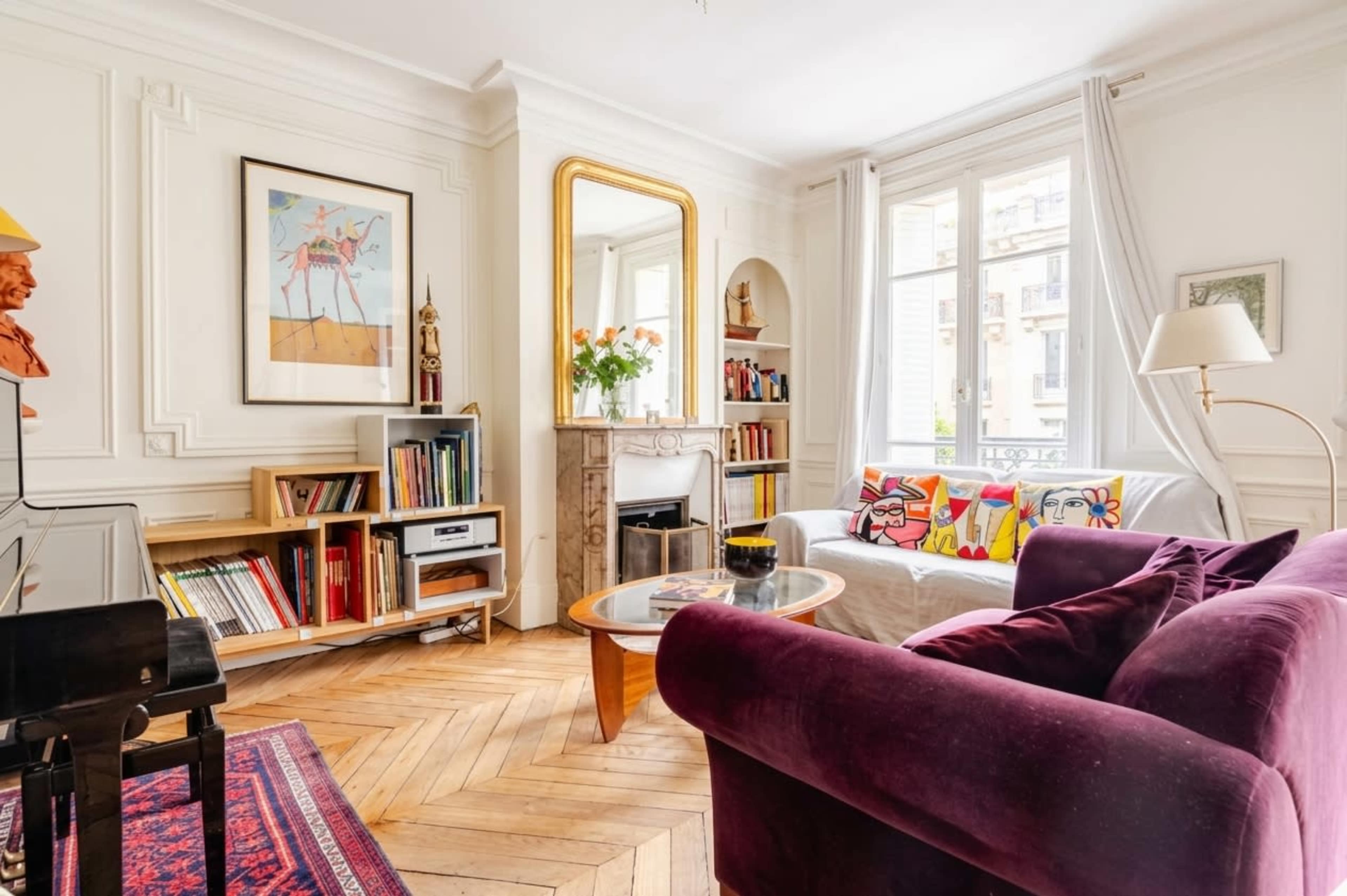 The image shows a stylish living room featuring a white sofa with colorful cushions, a purple armchair, bookshelves filled with books, and a large mirror above a decorative fireplace.