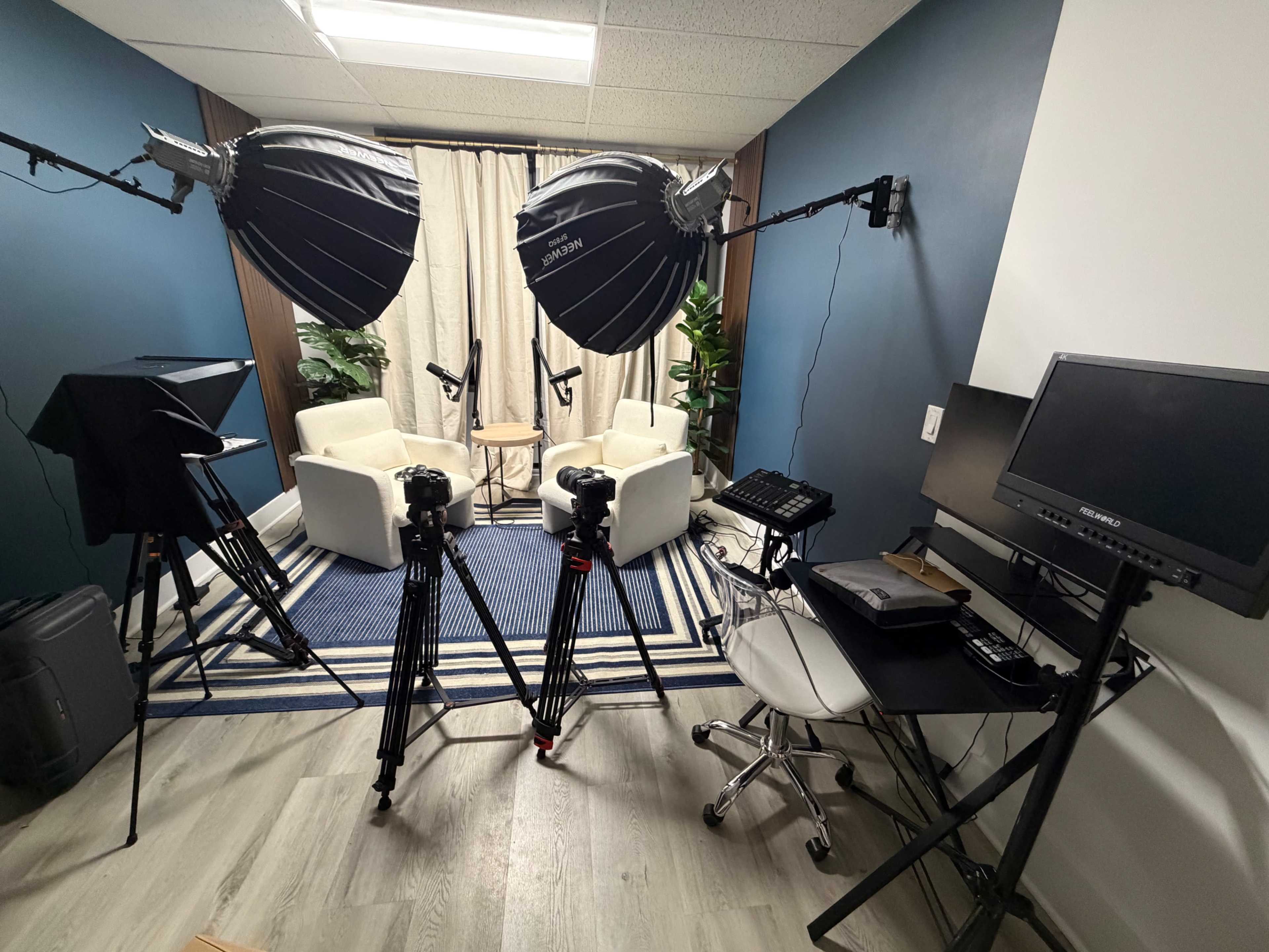 The image shows a small studio setup with two cameras on tripods, softbox lights, a table with two chairs, and a computer monitor against a blue wall.
