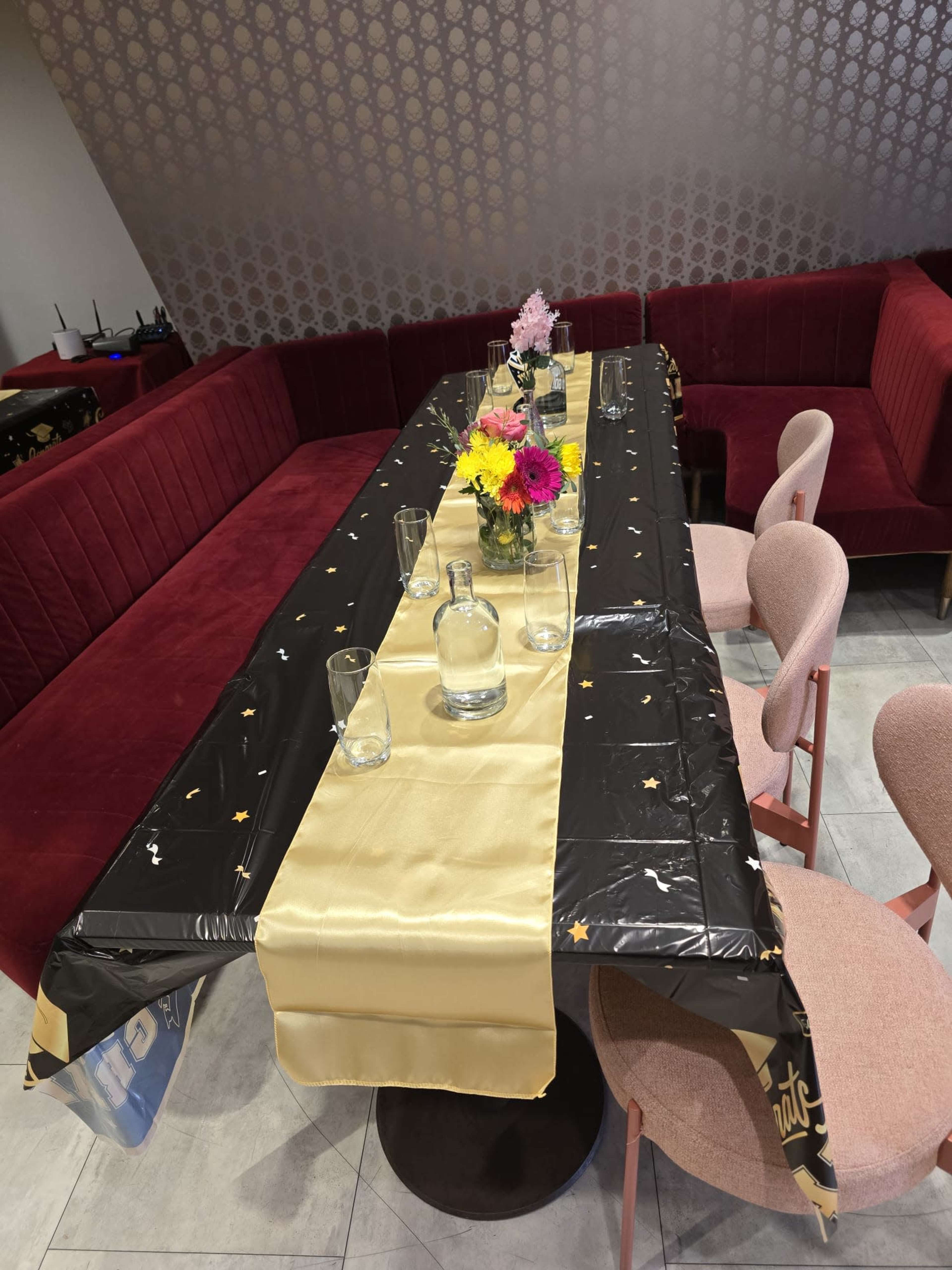 The image shows a long dining table covered with a black and gold tablecloth, set with glassware and a floral centerpiece, surrounded by pink chairs and a red upholstered seating area.