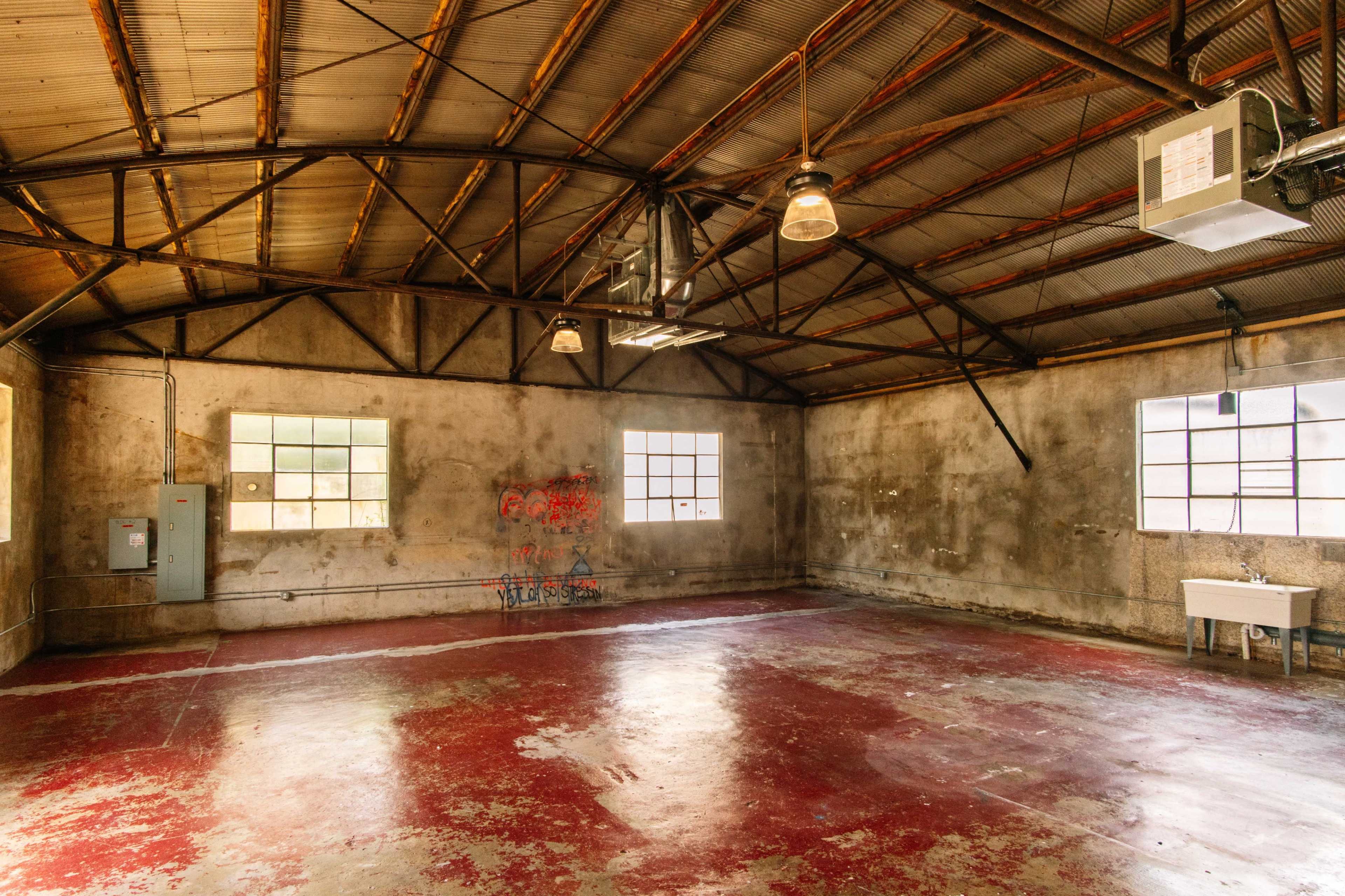 The image shows an empty industrial space with concrete walls, large windows, and a red-painted floor.