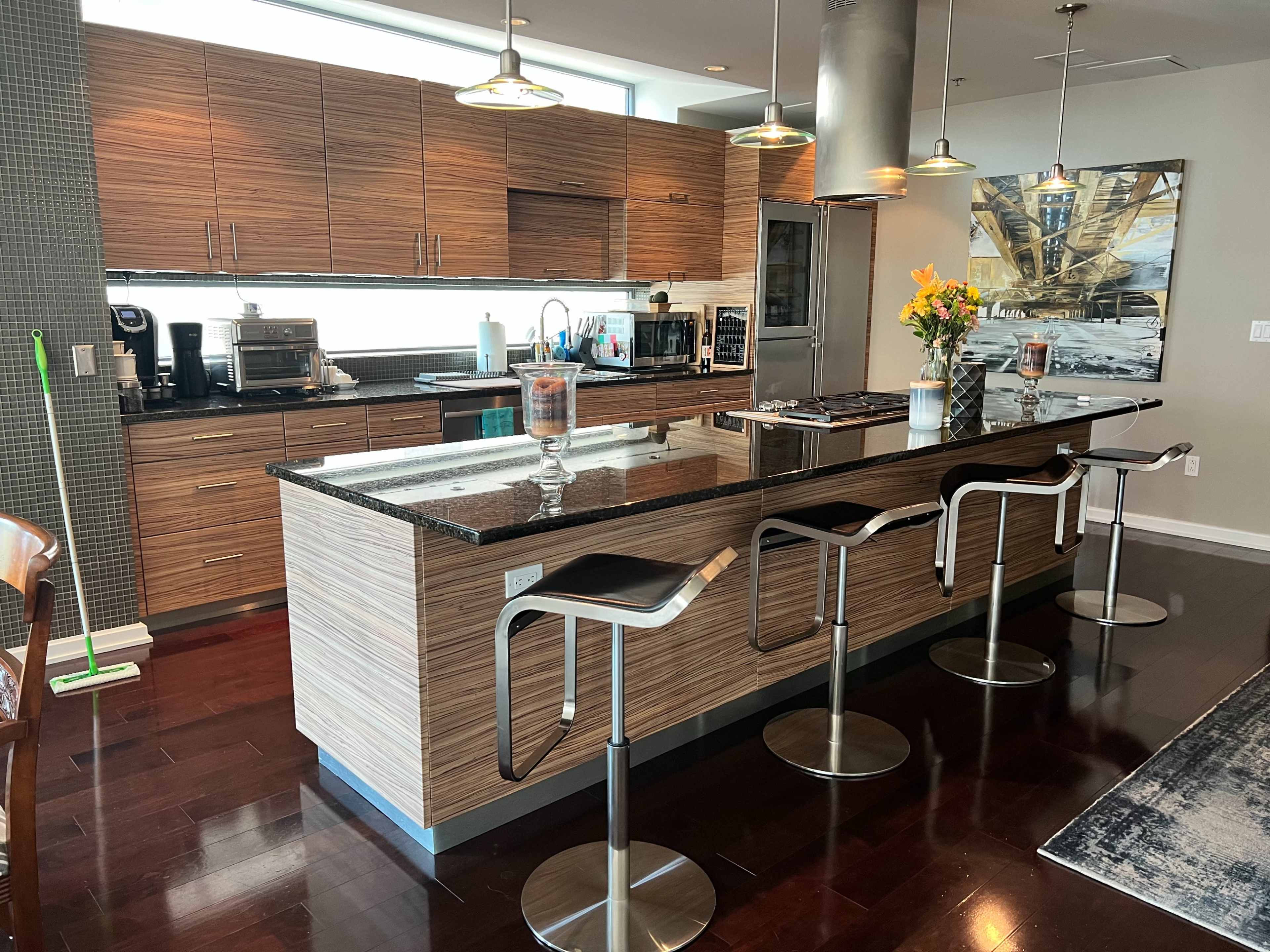 A modern kitchen with a central island, bar stools, and wooden cabinetry, featuring appliances and a vase of flowers.