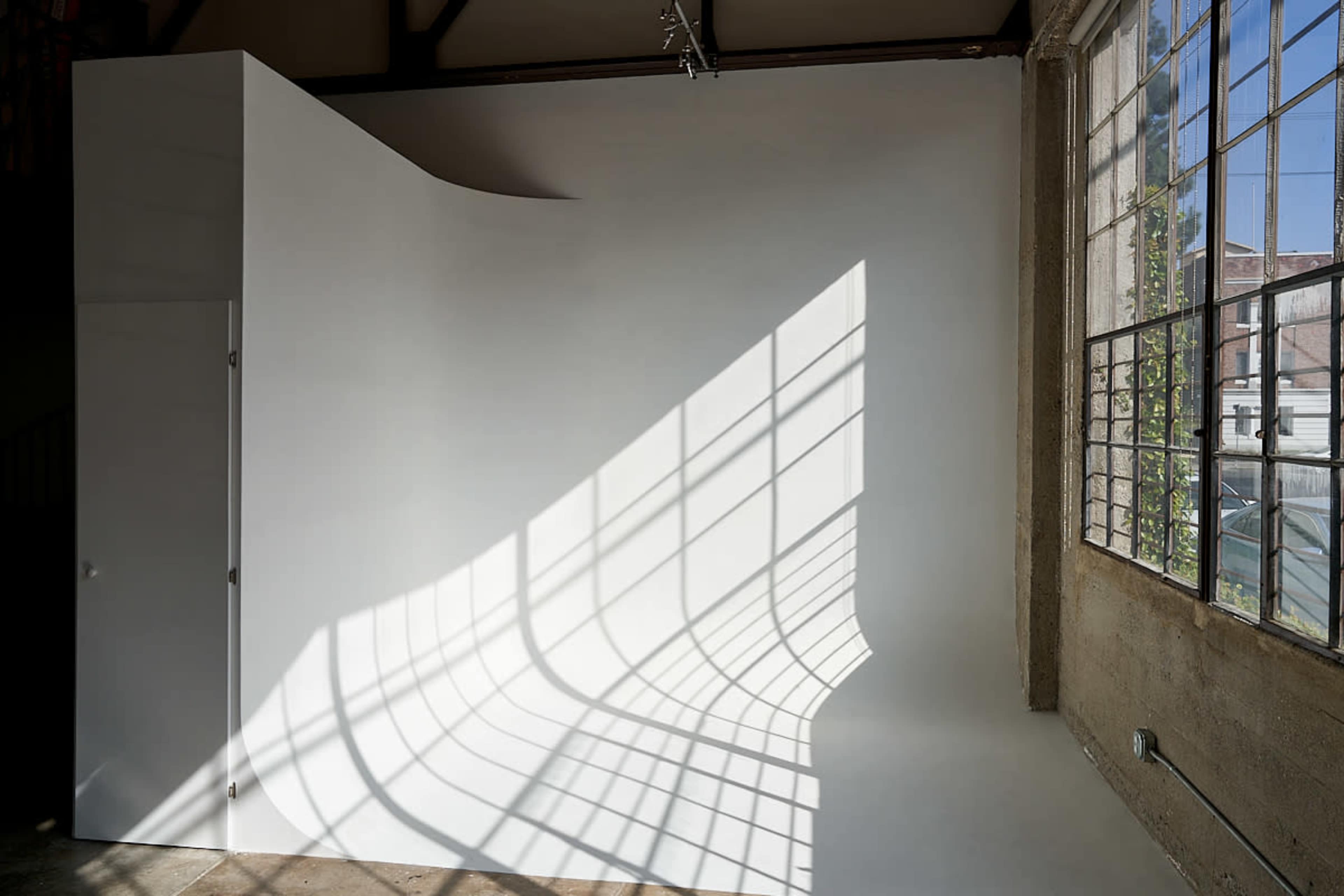 A minimalist studio space with a curved white wall and sunlight casting geometric shadows from a grid-patterned window.