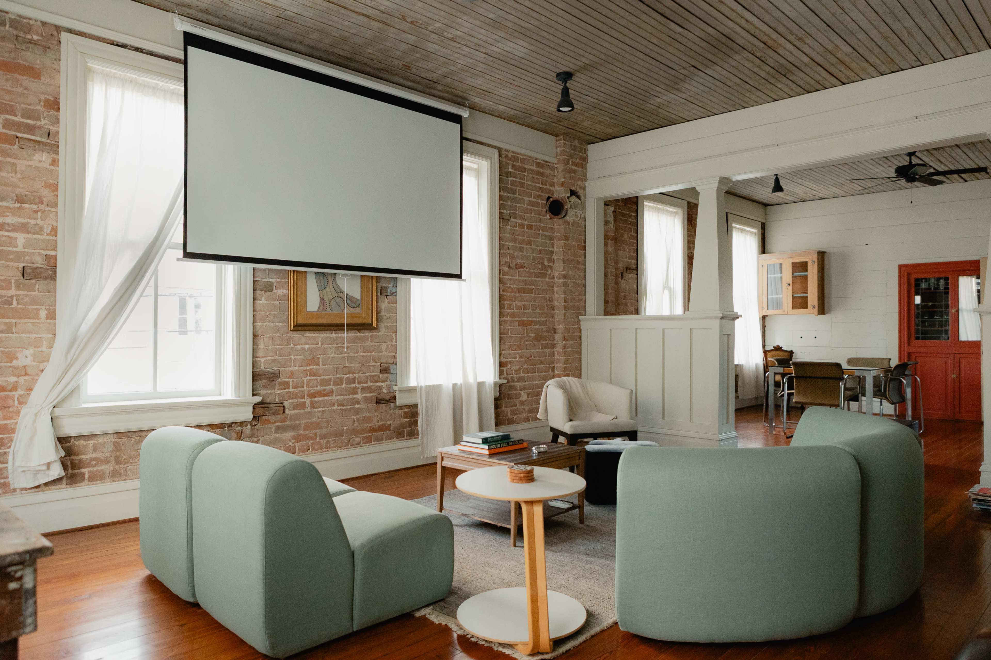 The image shows a cozy living room with a large projection screen, two light green sofas, and a small round coffee table, set against a backdrop of exposed brick walls and natural light from windows.
