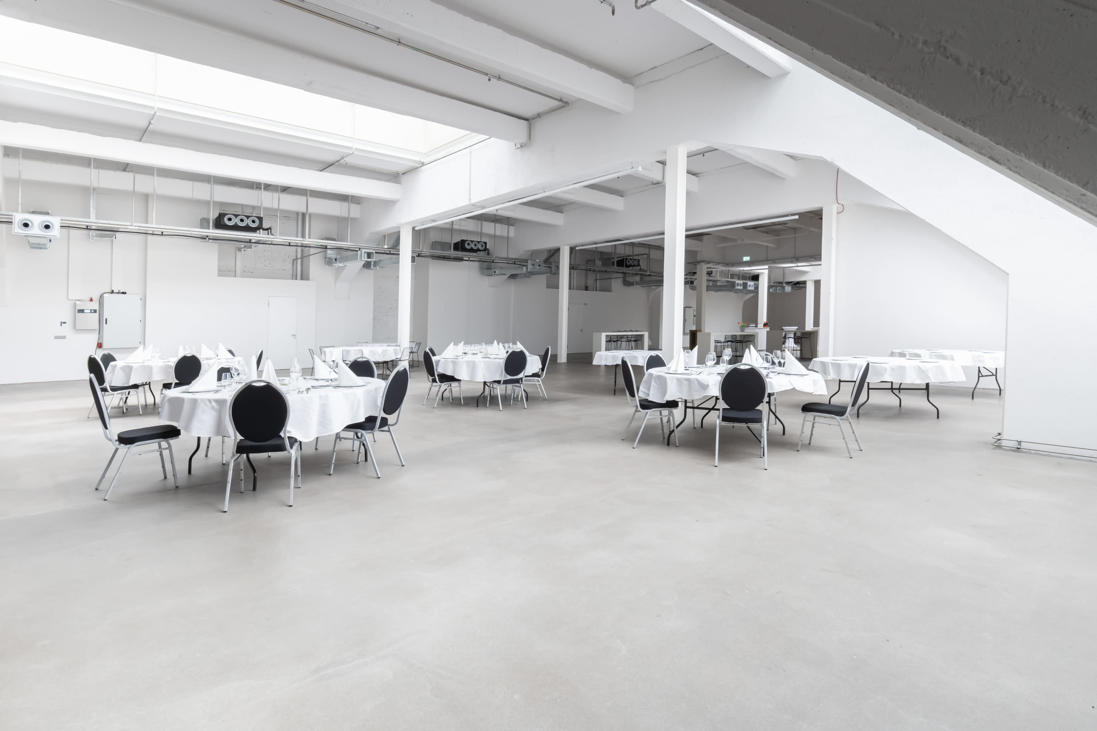 A large, airy event space features several round tables set with white tablecloths and black chairs arranged throughout the room.
