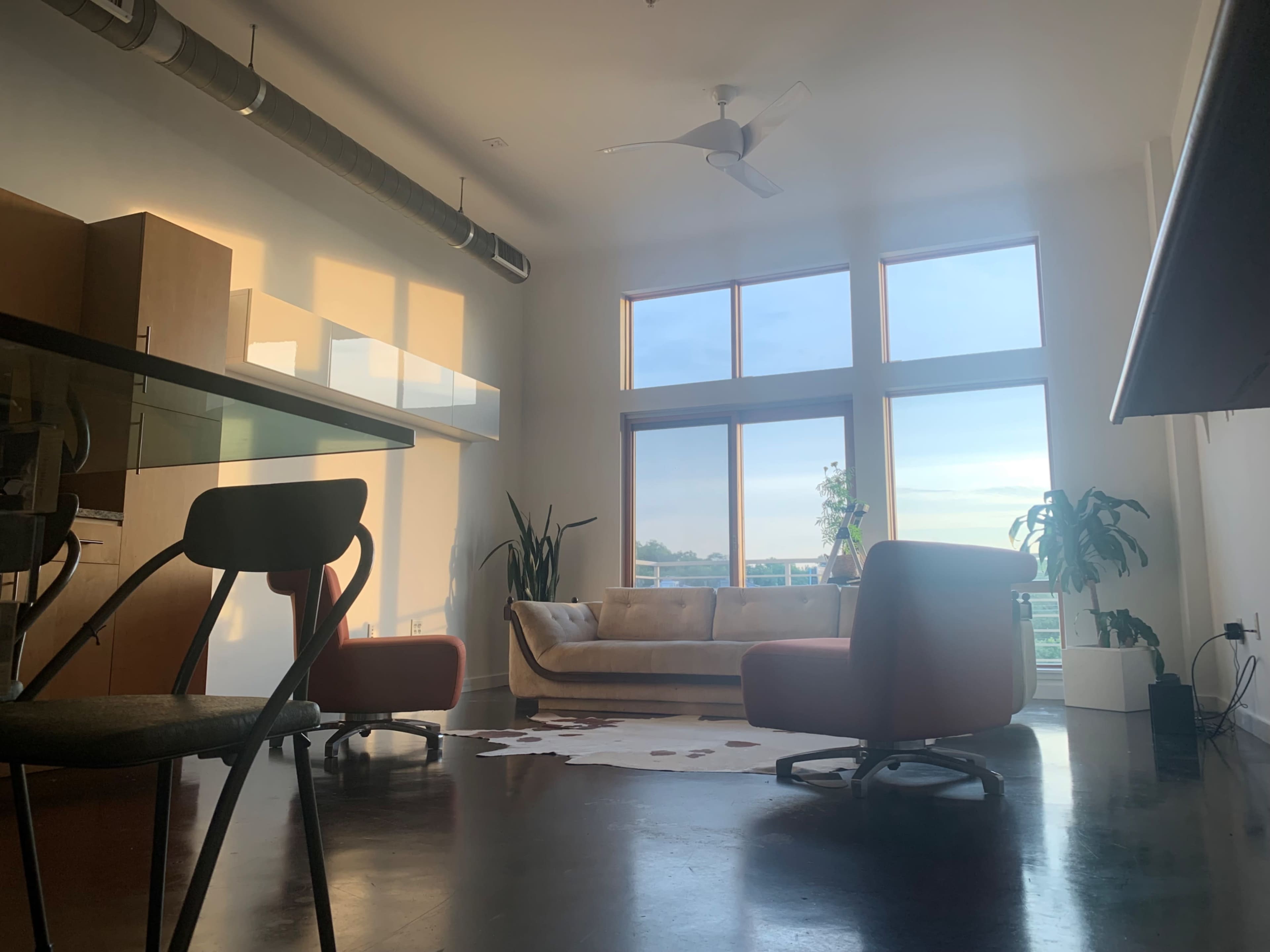 The image depicts a modern living room with large windows, a neutral sofa, and two orange chairs, all set over a dark polished floor.