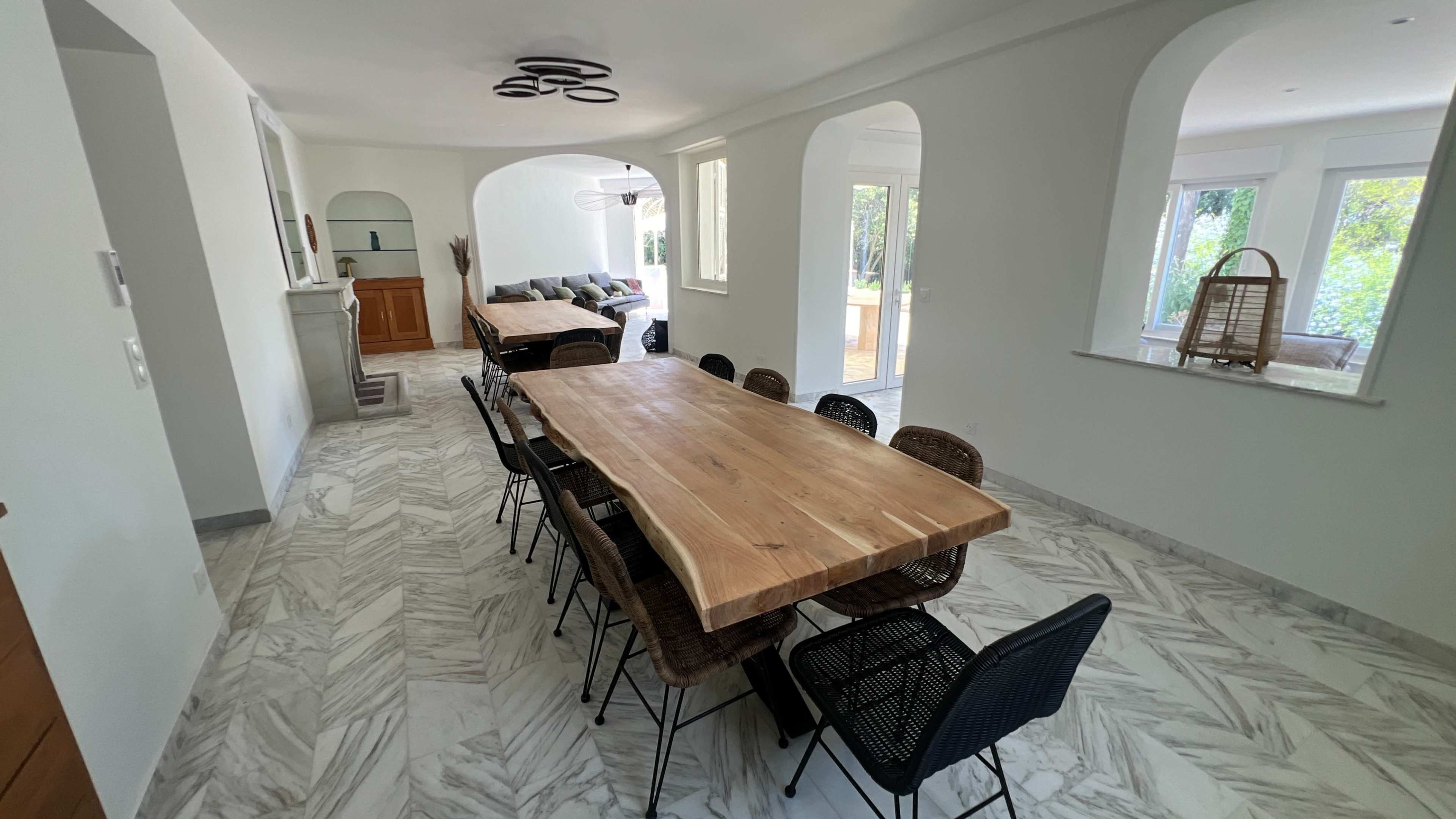 The image shows a spacious dining area with a large wooden table surrounded by black chairs, leading into a well-lit living space with large windows.