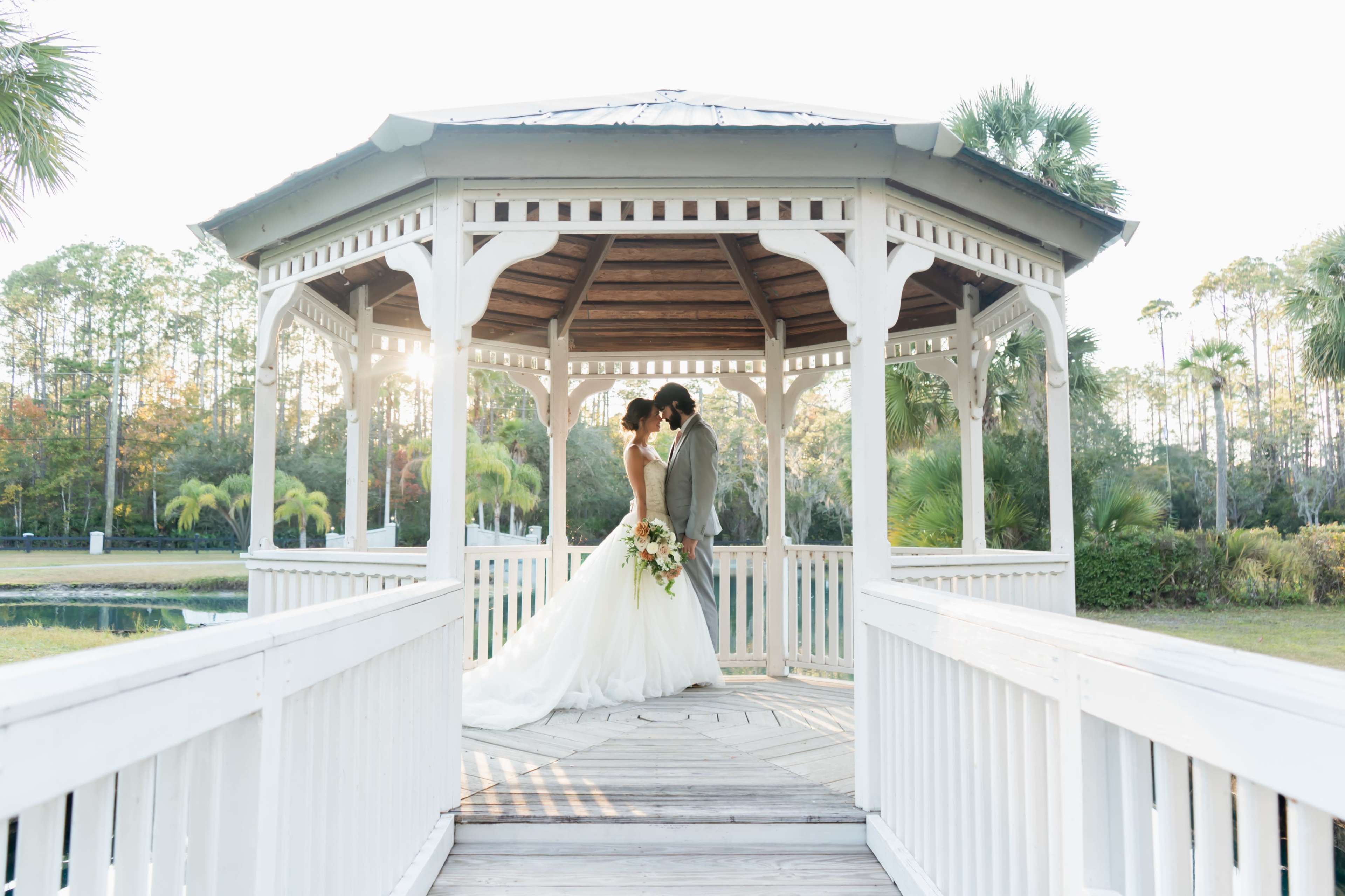 A couple in wedding attire shares a kiss under a gazebo surrounded by lush greenery.