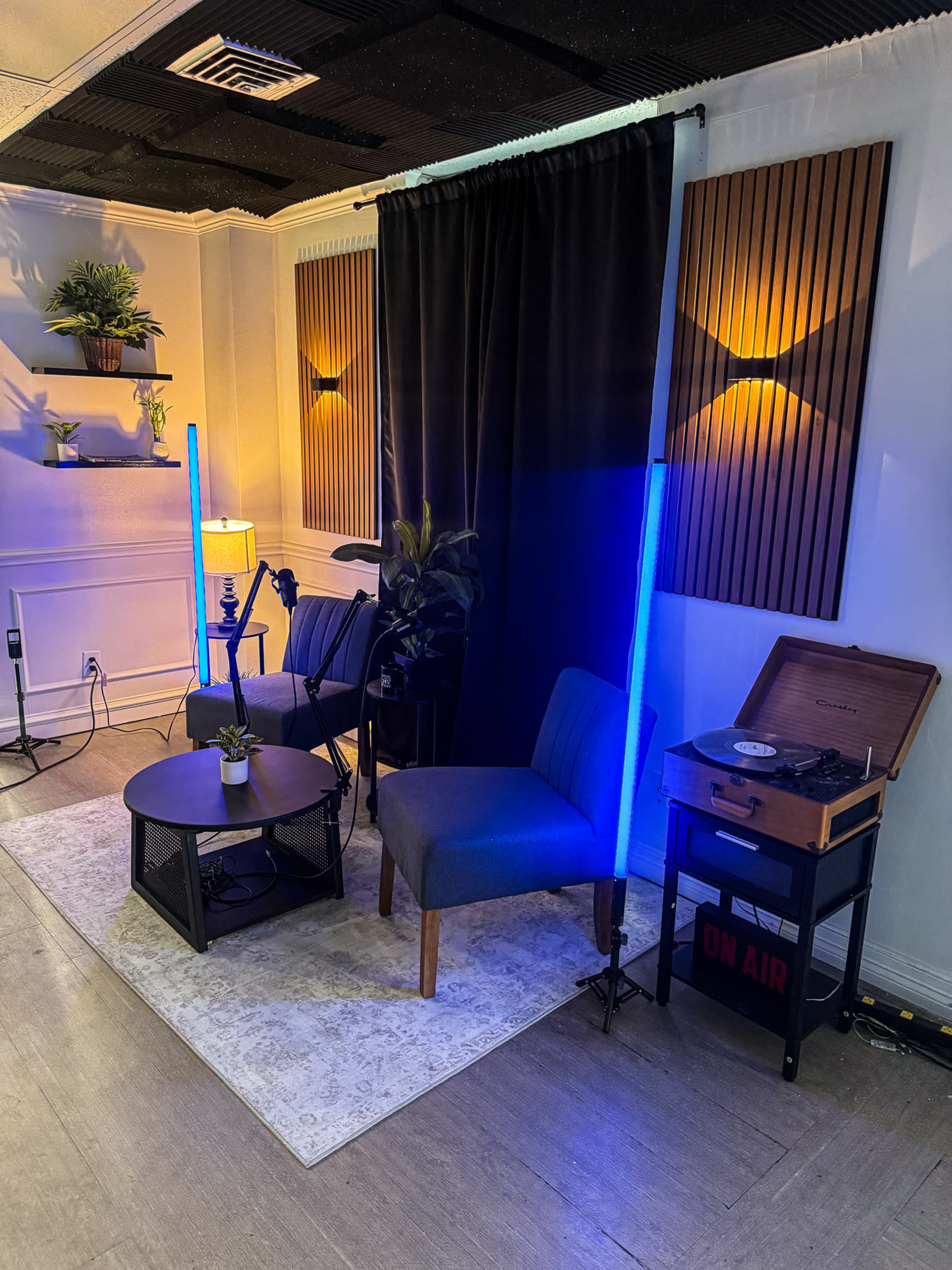 The image shows a cozy recording setup featuring a small table and chairs, decorative plants, a standing lamp, and a vintage record player with an "ON AIR" sign.