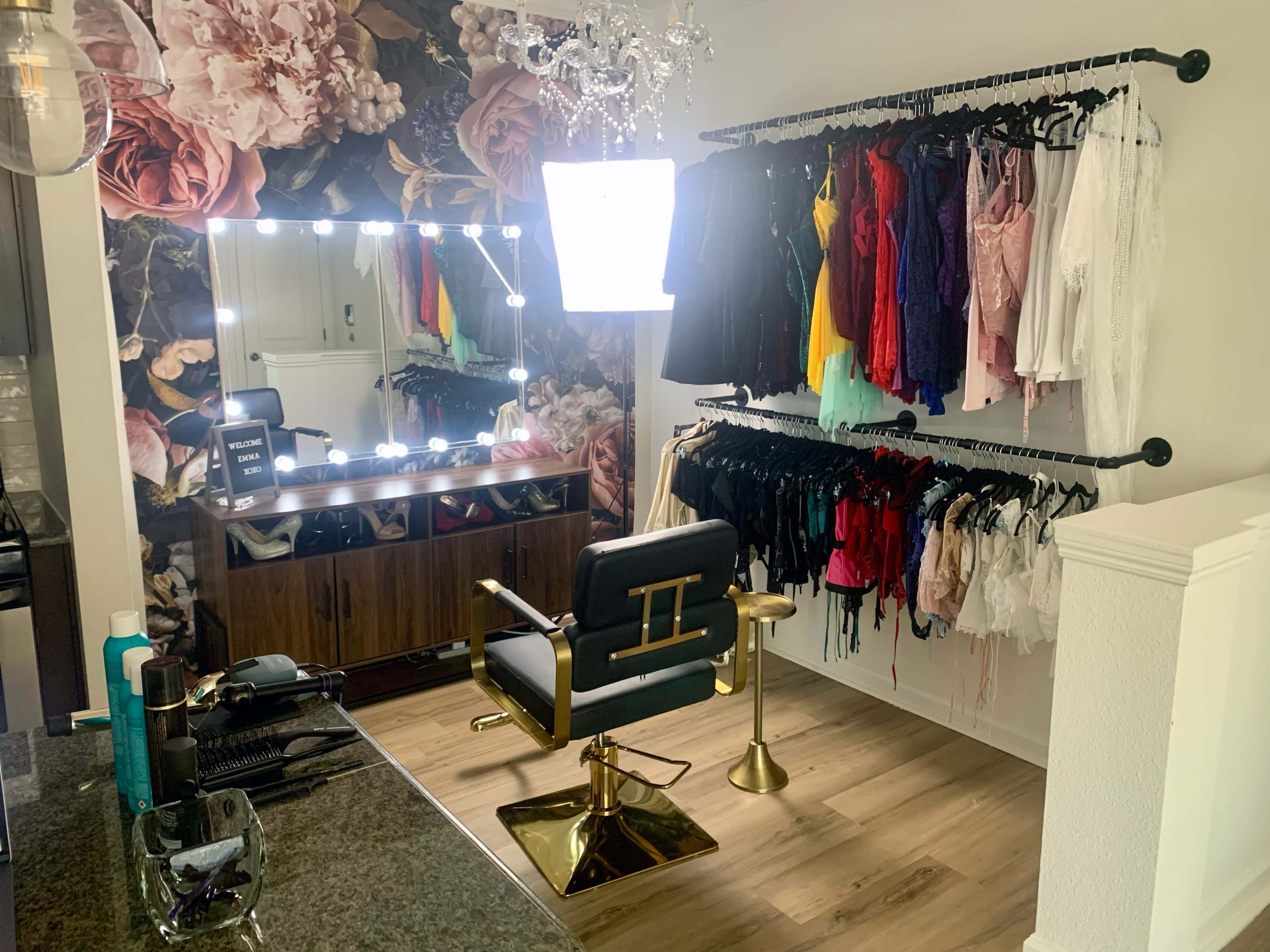 The image shows a styled dressing area featuring a makeup station with a large illuminated mirror, surrounded by a variety of colorful clothing hanging on racks.
