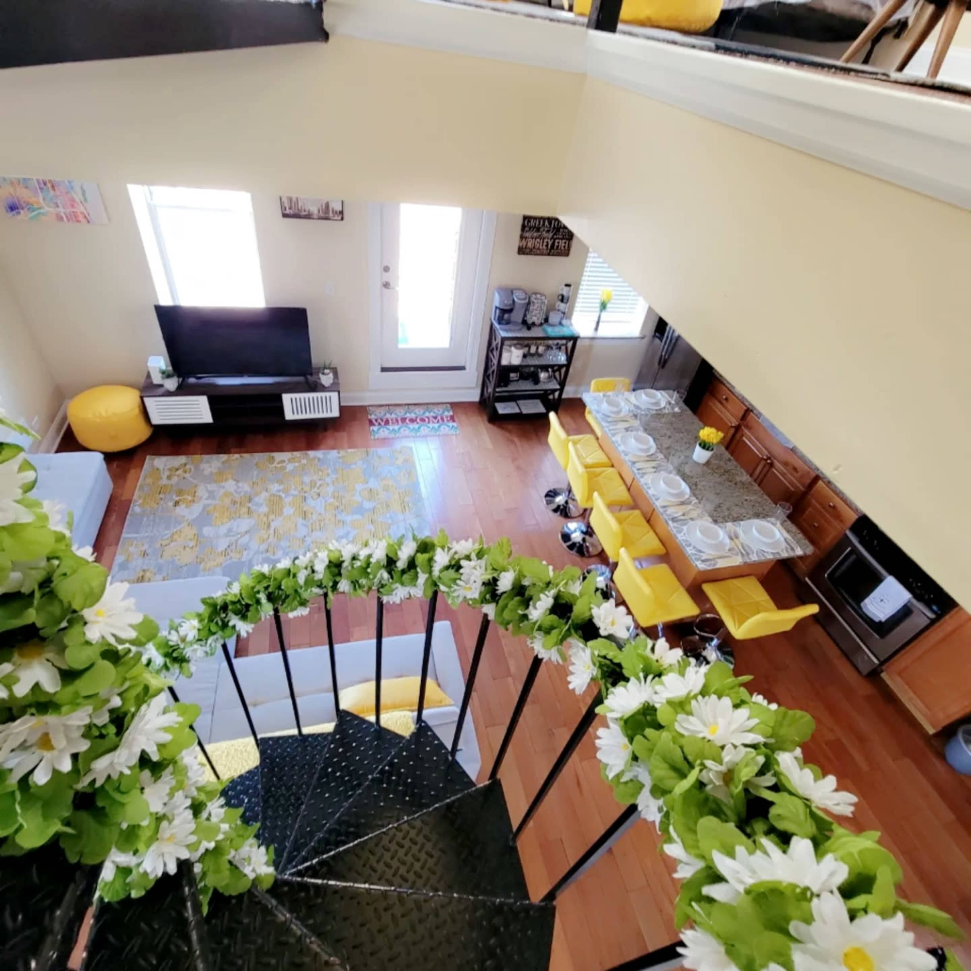 A spiral staircase with a floral decoration leading down to a living area featuring a television, a dining table with yellow chairs, and a kitchen.