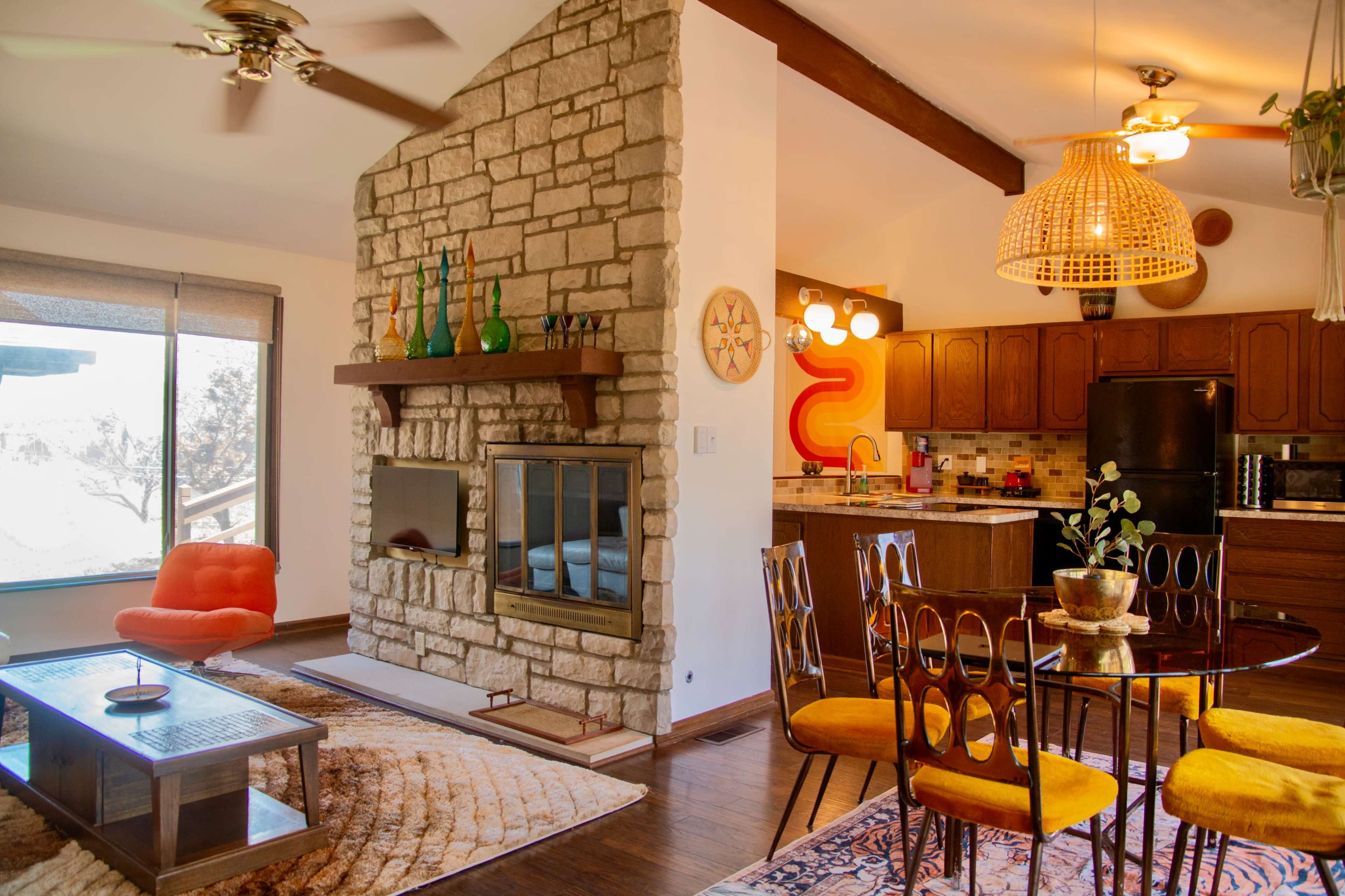 The image depicts a living room and kitchen area featuring a stone fireplace, a round dining table with orange chairs, and large windows that allow natural light to enter.