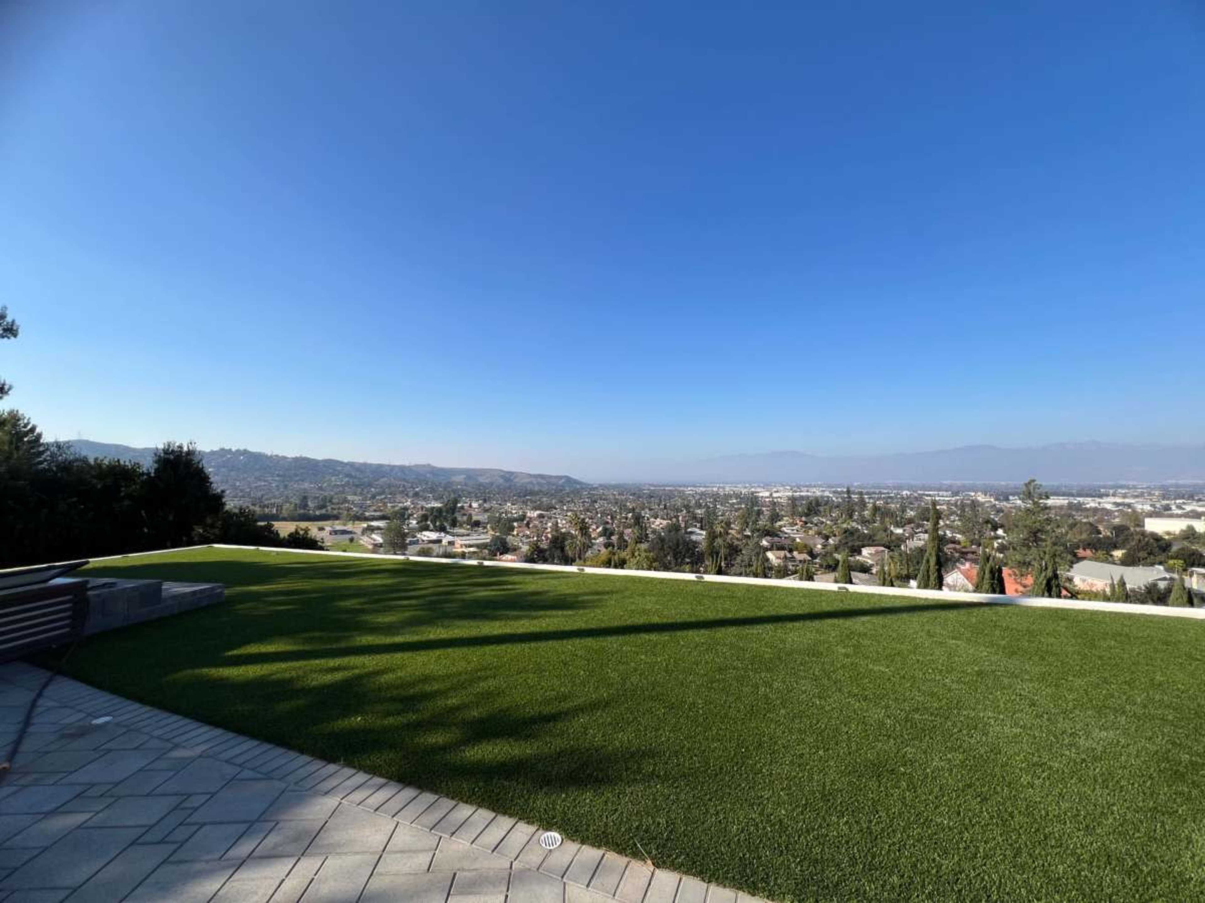 The image shows a flat, grassy area overlooking a suburban landscape and mountains under a clear blue sky.