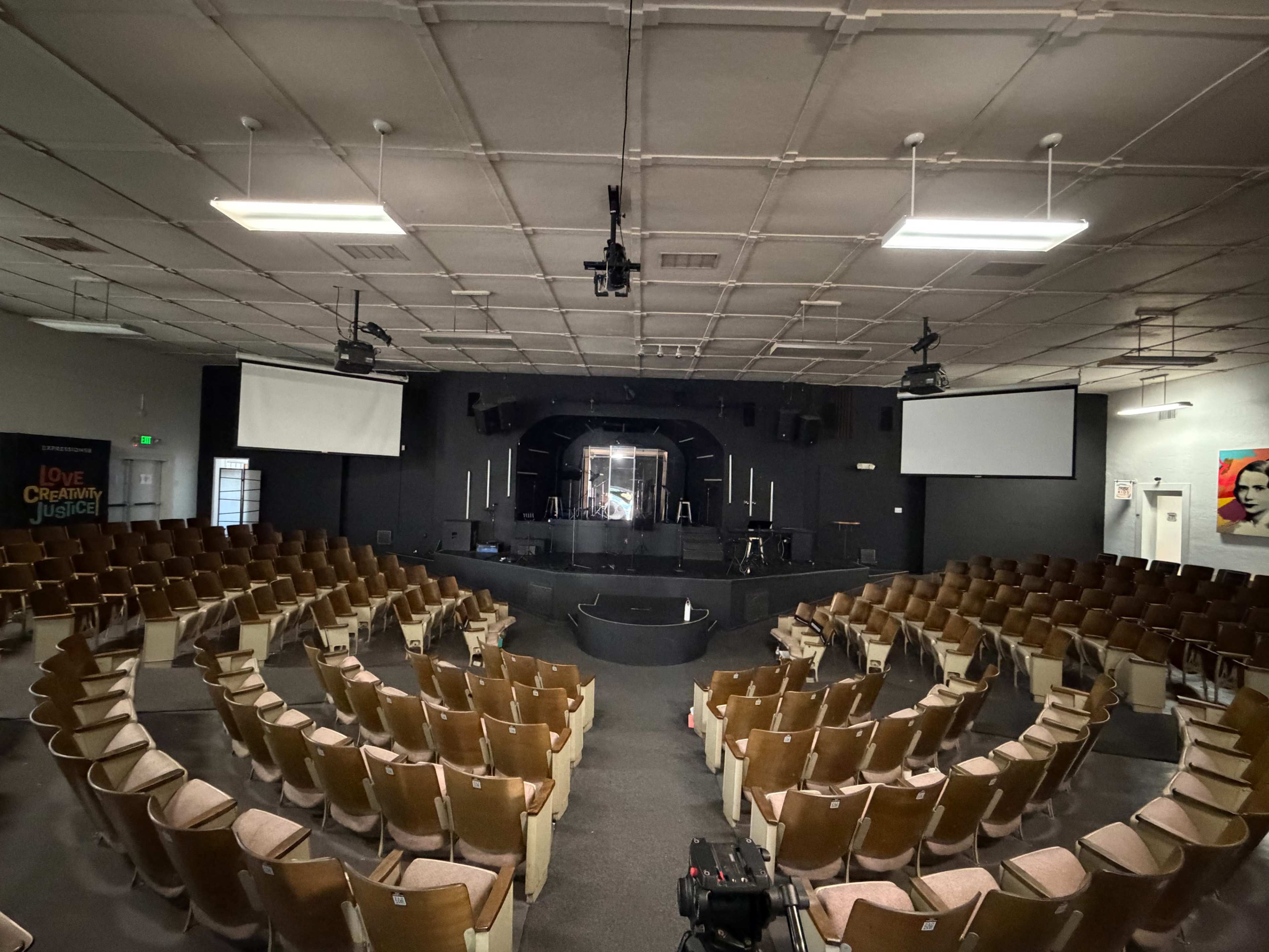 The image shows an empty auditorium with rows of wooden chairs facing a central stage, equipped with lighting and projection screens.