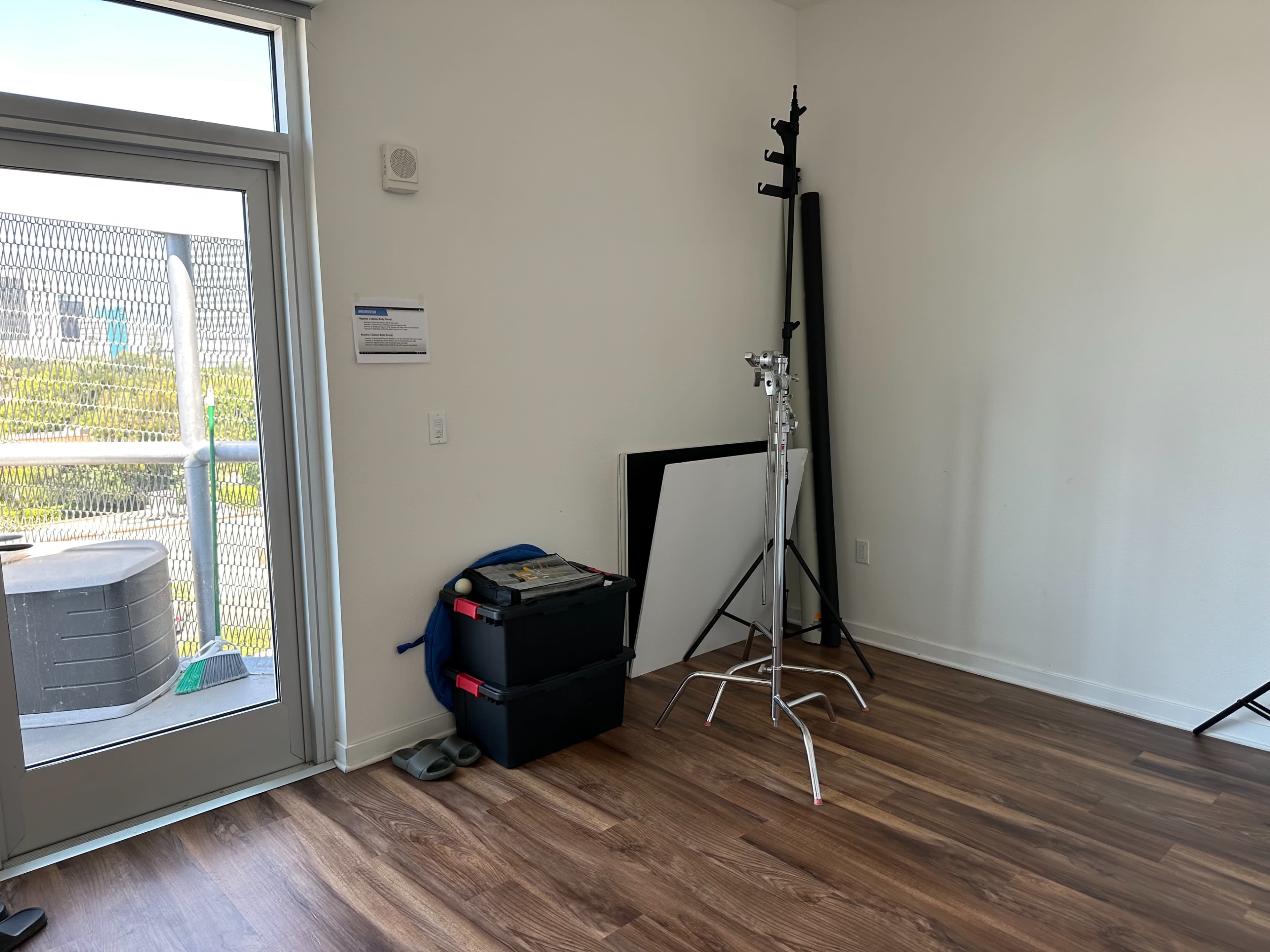 The image shows a sparsely furnished room with wooden flooring, a large window, a foldable backdrop stand, storage boxes, and a few equipment stands.